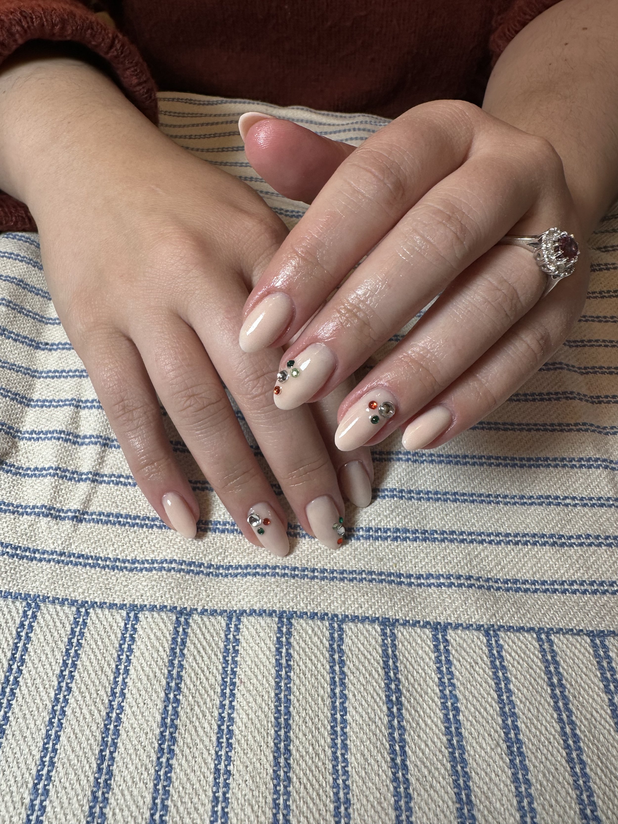 Close-up of hands with manicured nails, some decorated with small colored rhinestones, resting on striped fabric. One hand wears a ring with a red gemstone surrounded by diamonds.