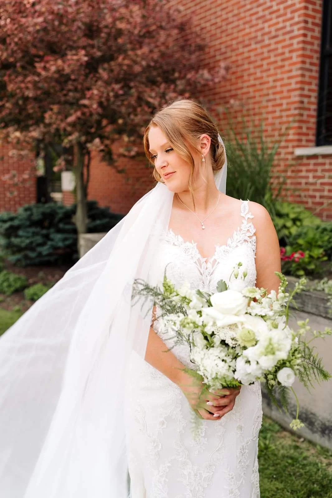 A bride in a white lace wedding dress with a veil, holding a bouquet of white flowers, standing outdoors near a brick building and greenery.