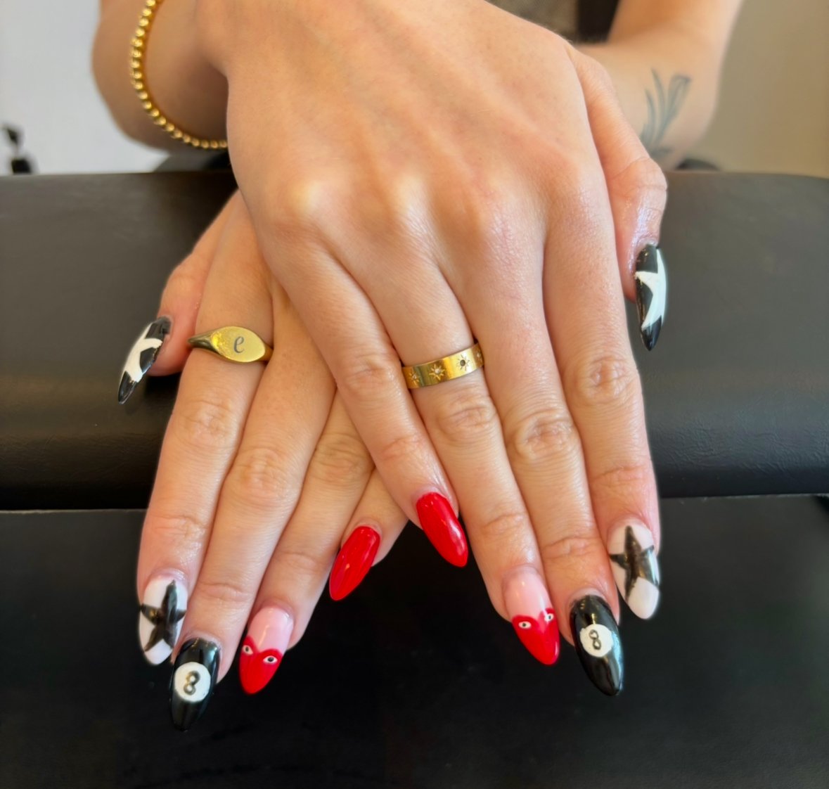 Close-up of two hands with decorated nails, wearing rings, one with a gold bracelet, resting on a black surface.