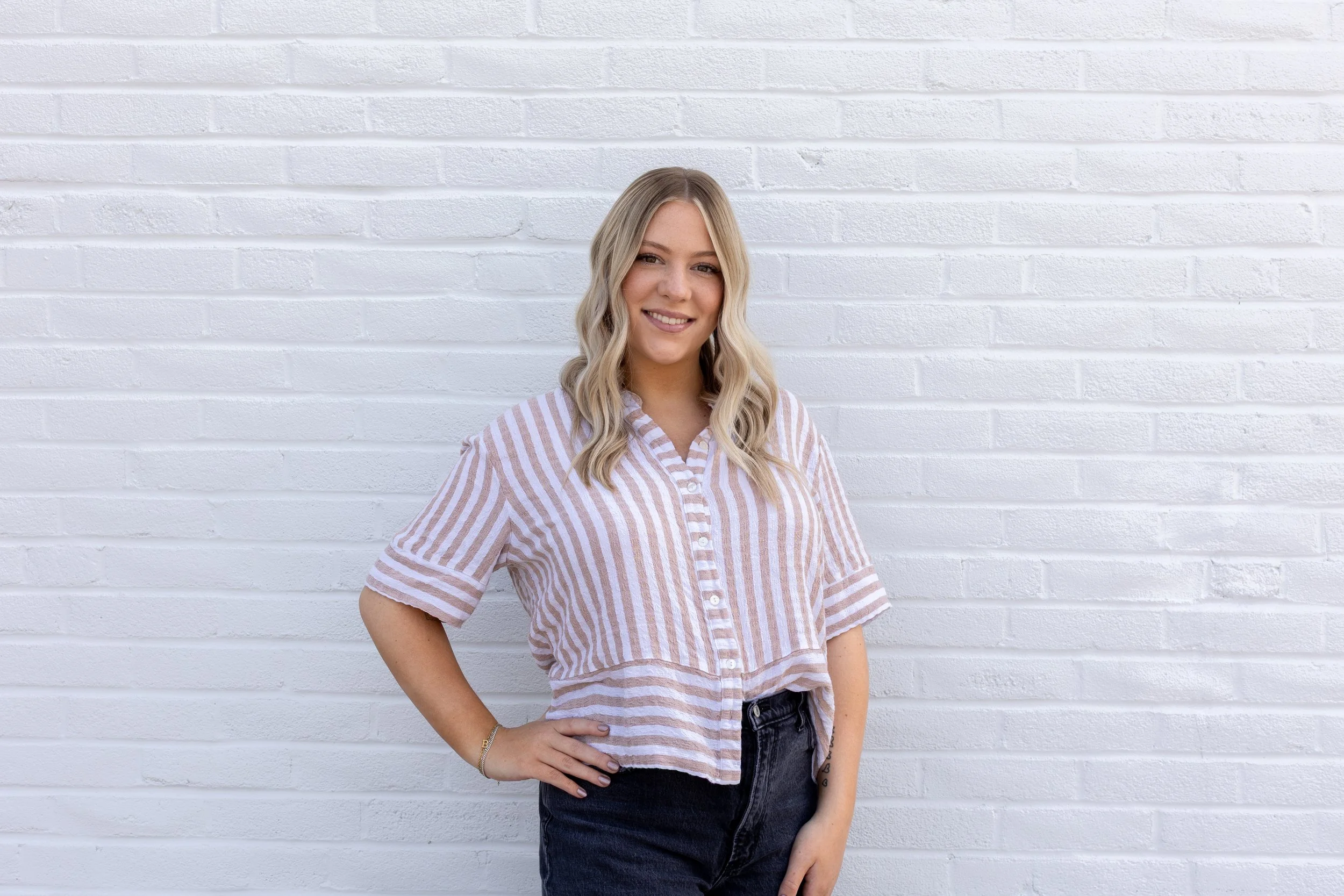 A young woman with blonde wavy hair smiling and standing against a white brick wall, wearing a pink and white striped button-up shirt and black jeans.