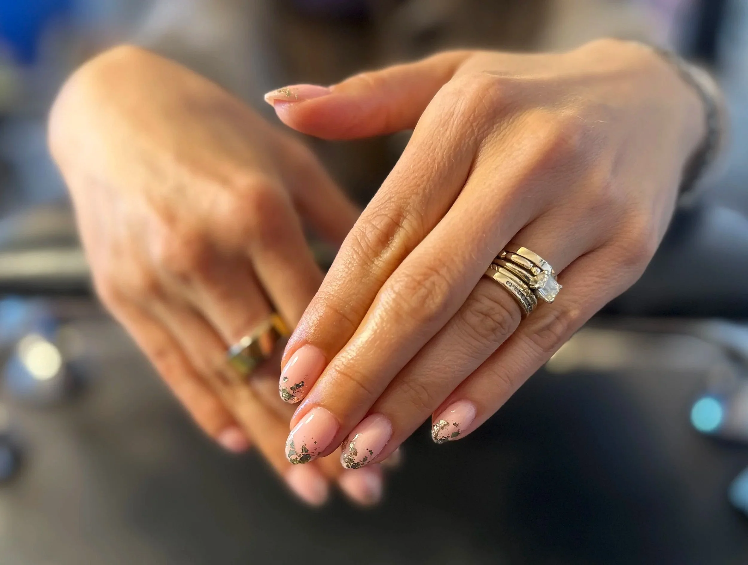 Close-up of two hands with decorated nails, wearing rings, one with a gold bracelet, resting on a black surface.