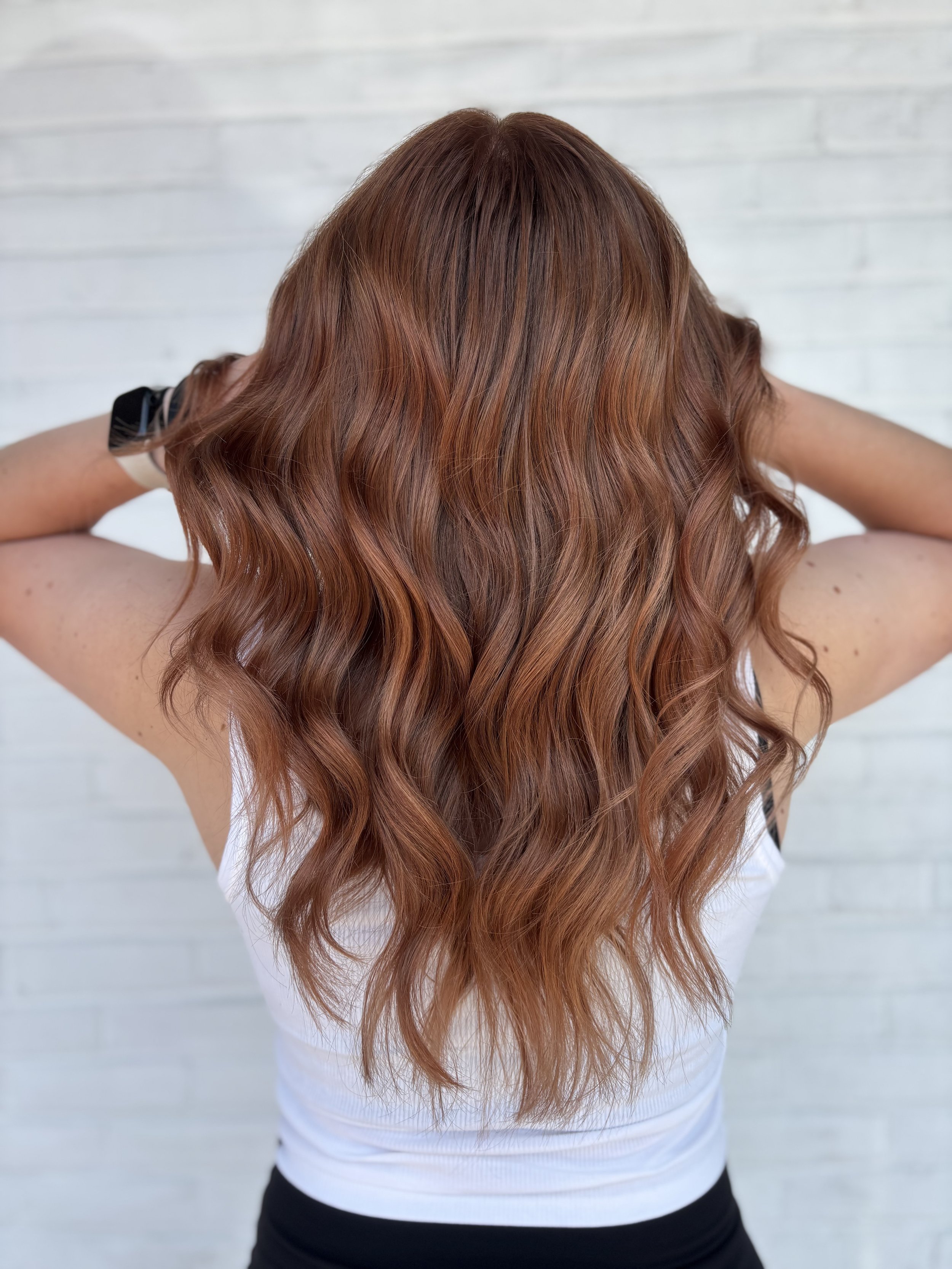 Back view of a woman with long, wavy, auburn hair wearing a white tank top against a white brick wall.