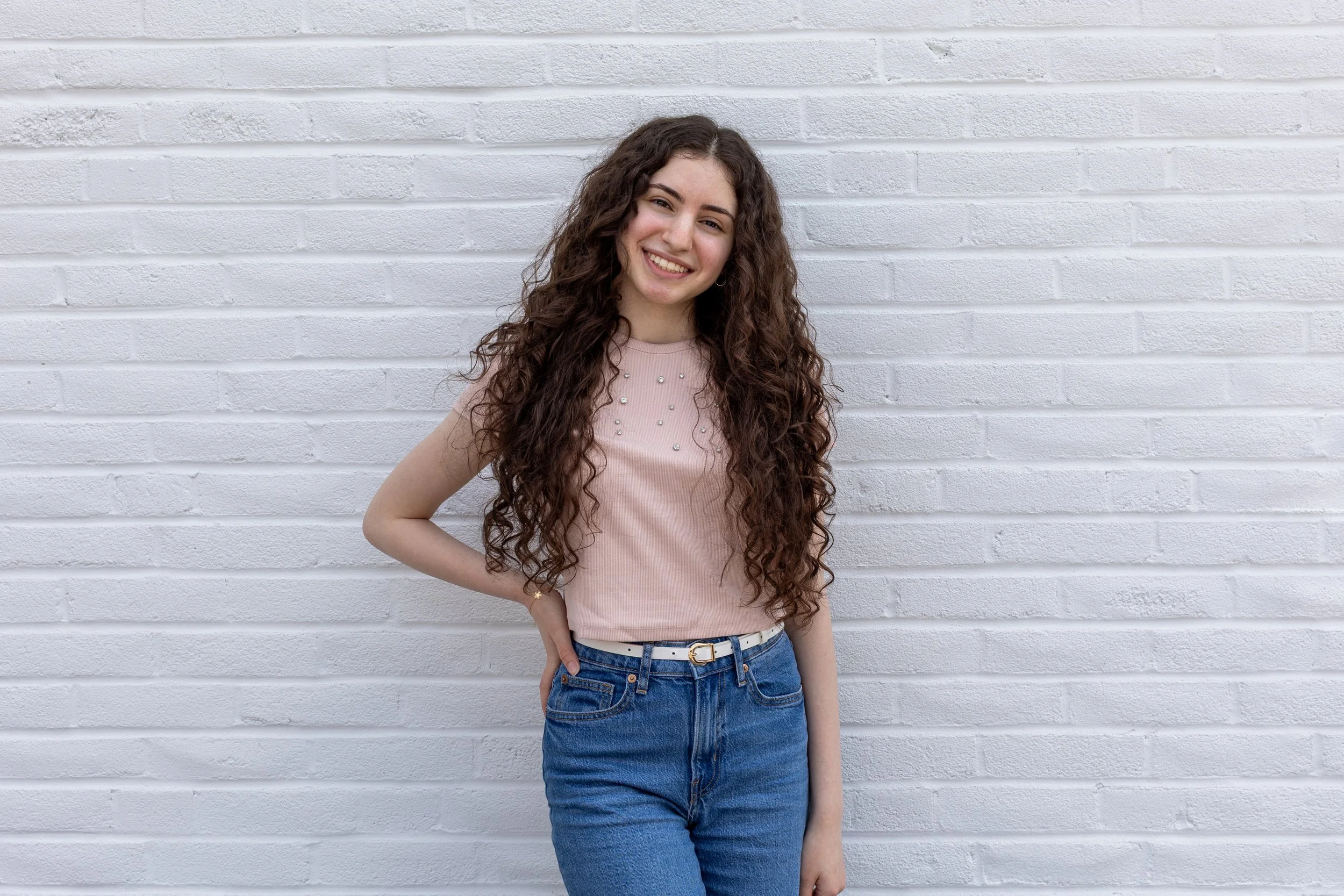 A young woman with long curly hair smiling and standing against a white brick wall, wearing a pink top with pearl embellishments and blue jeans.