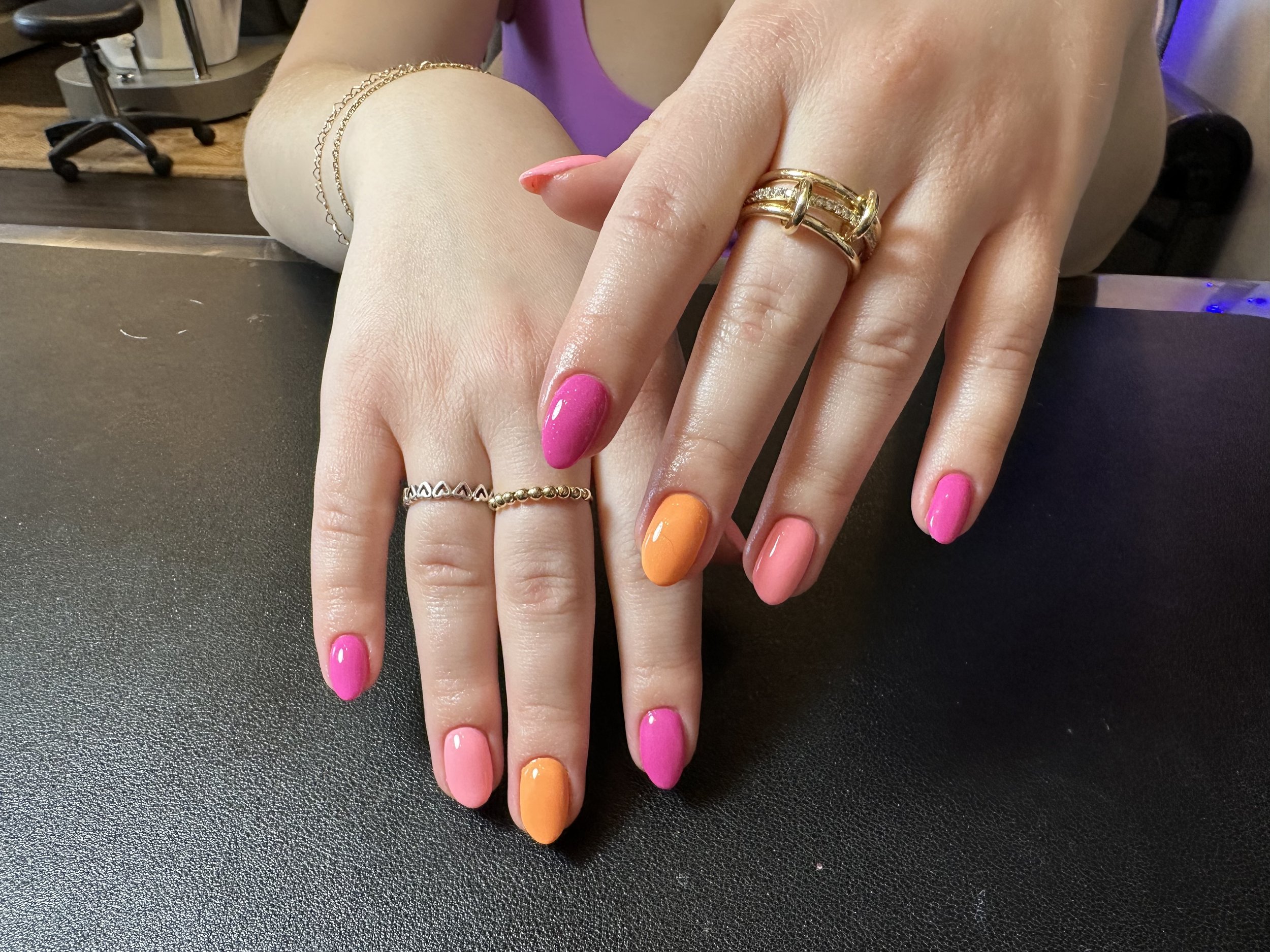 Close-up of a hand with colorful painted nails, gold rings, and gold bracelets resting on a dark surface, with a blurred background showing a person wearing a purple top.