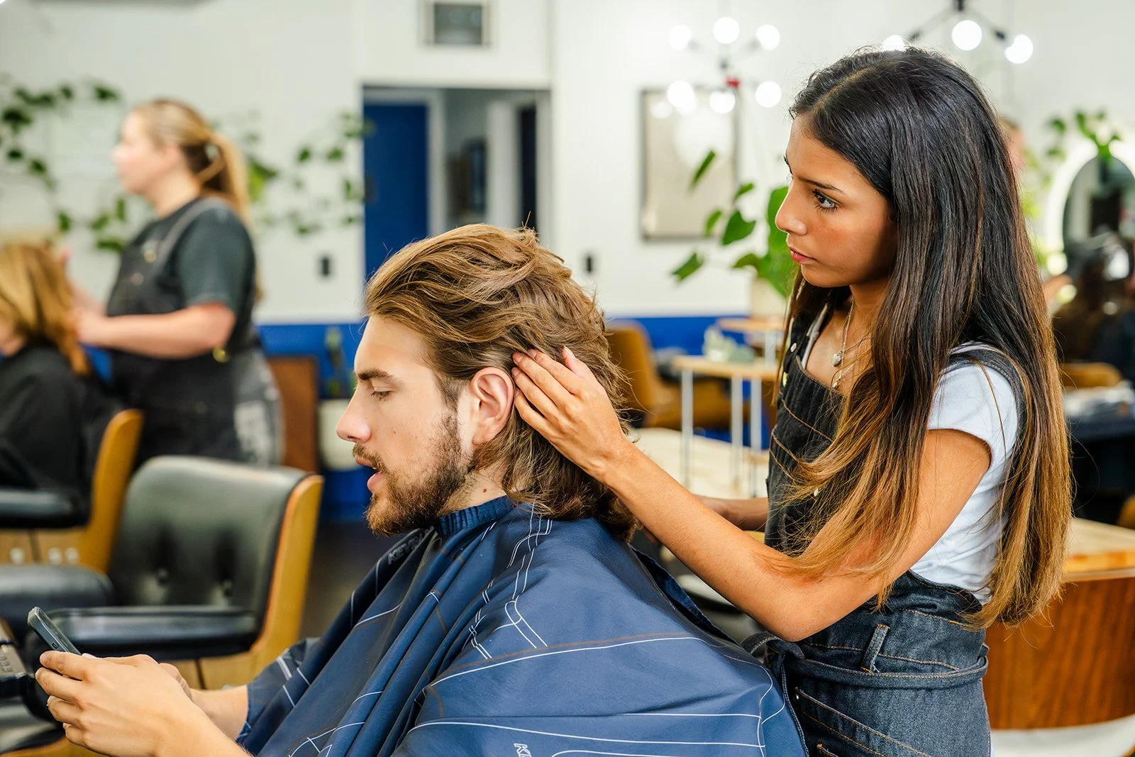 A woman is giving a man a head massage in a hair salon. The man is sitting with his eyes closed, holding a phone, while the woman gently touches his head.