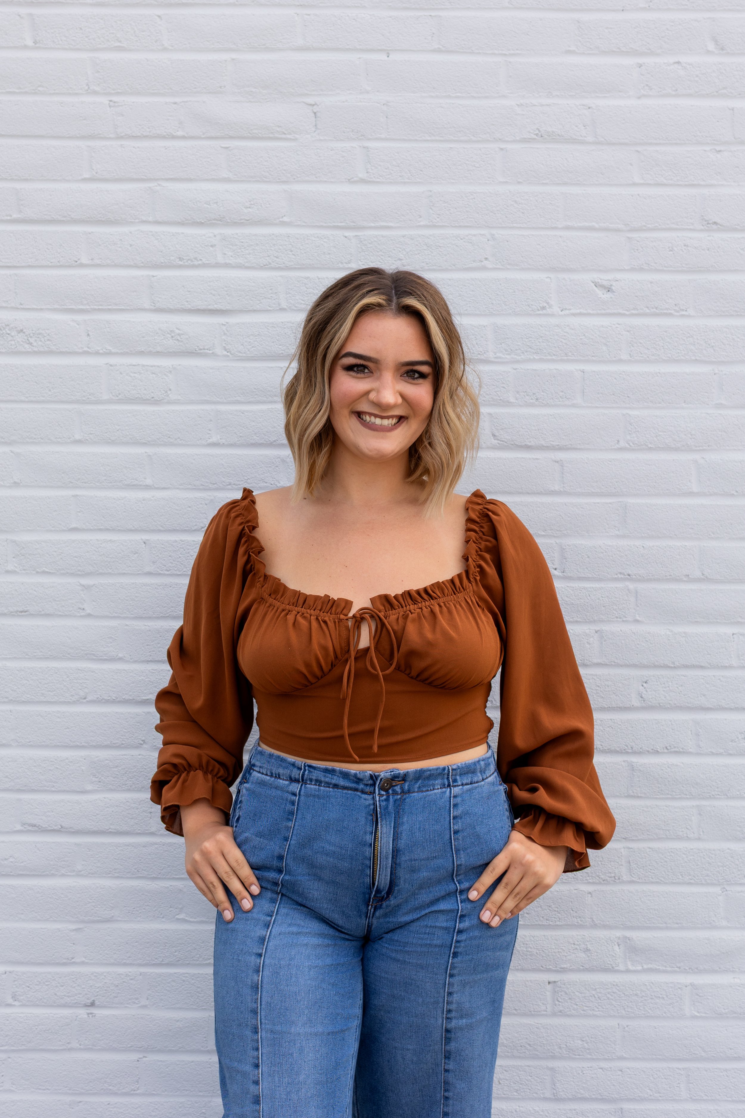 A young woman with blonde hair and a big smile, wearing a brown blouse with ruffled sleeves and high-waisted blue jeans, standing against a white brick wall.