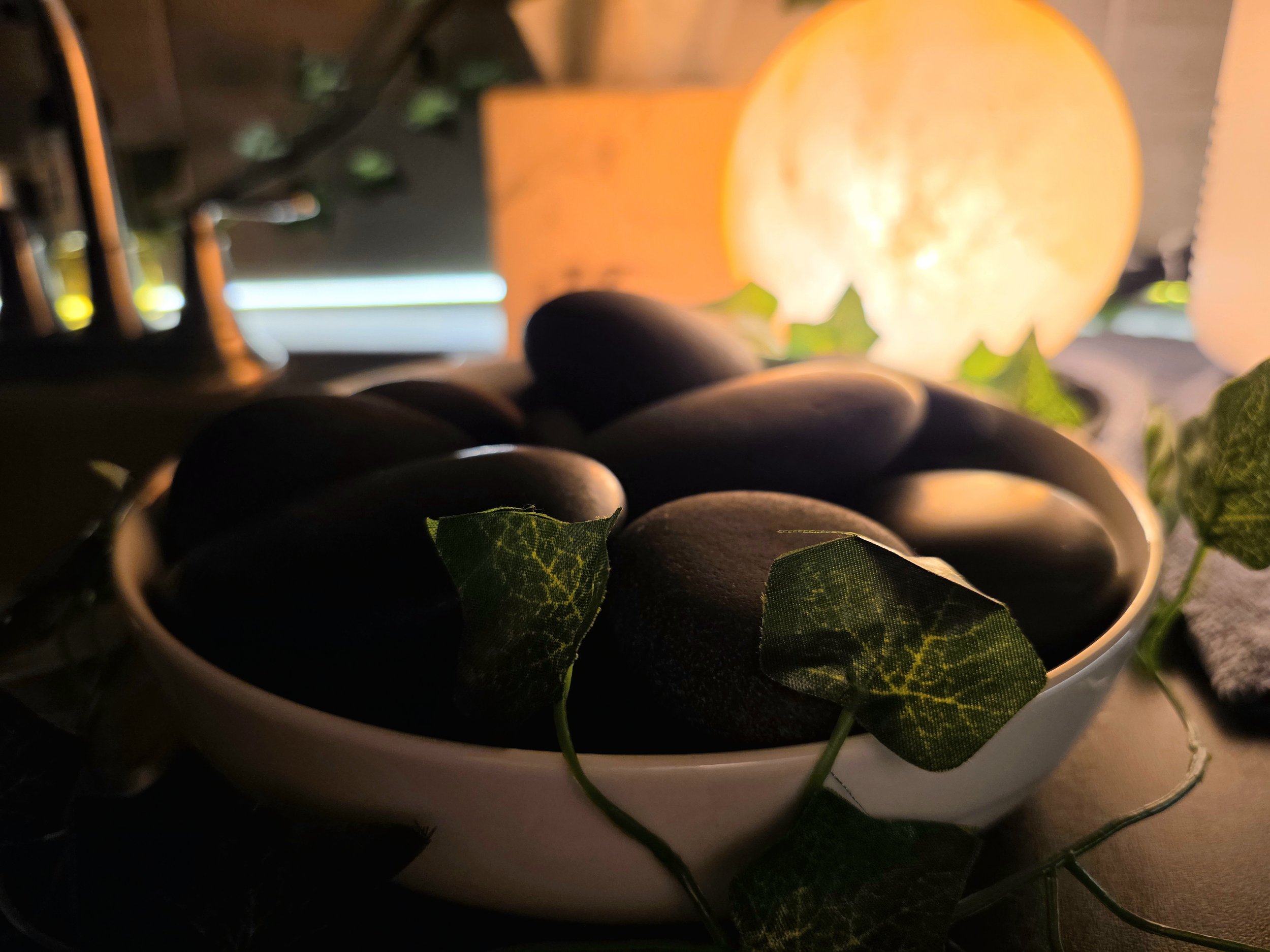 A bowl filled with black stones and green ivy leaves, with a glowing orange pumpkin in the background.