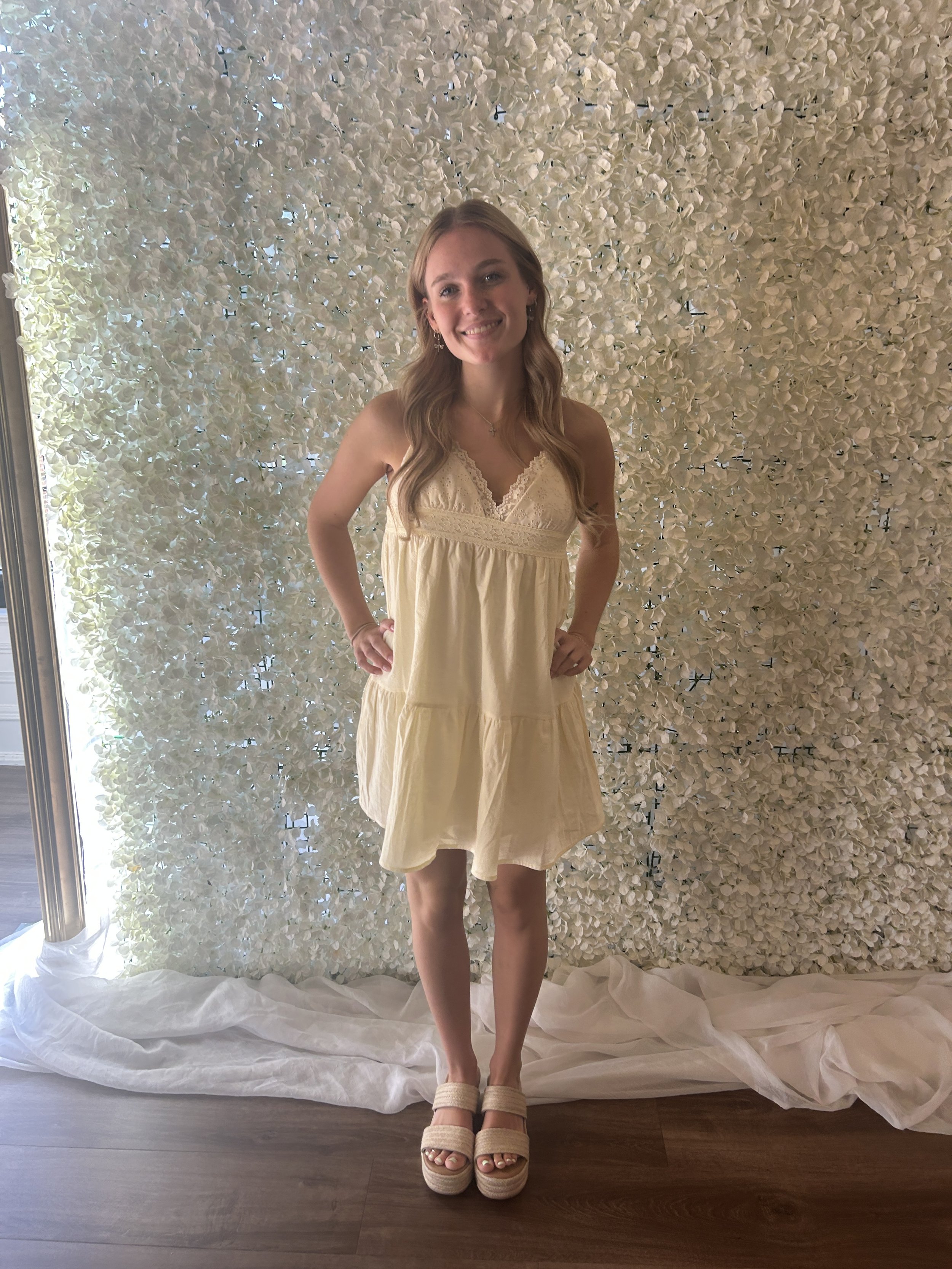 A young woman with long wavy hair wearing a light yellow sundress with lace details, standing in front of a backdrop made of white flowers. She is smiling with her hands on her hips and wearing beige platform sandals.