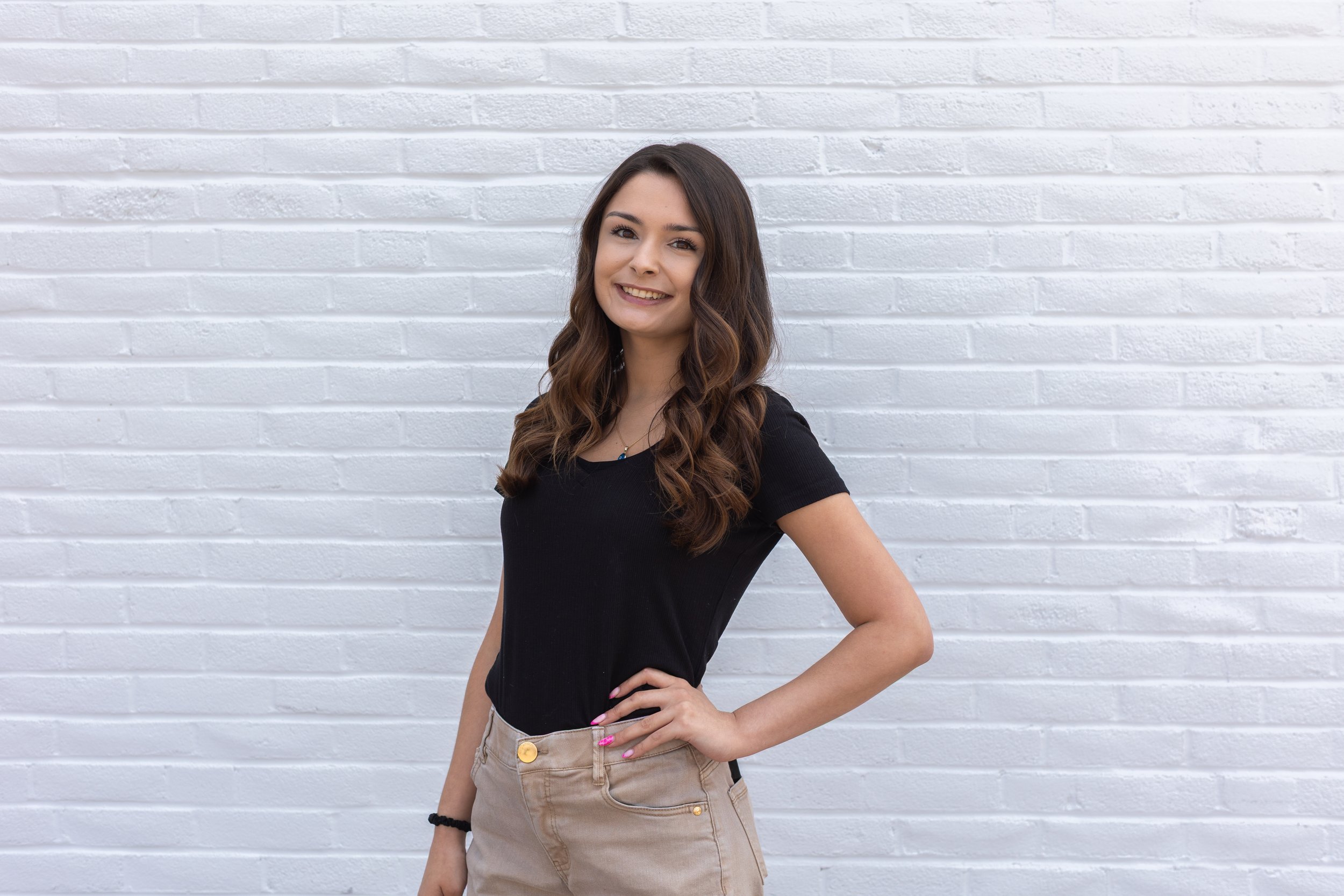 A young woman with long, wavy brown hair and a bright smile stands against a white brick wall. She is wearing a black short-sleeved top and beige shorts, with one hand on her hip.
