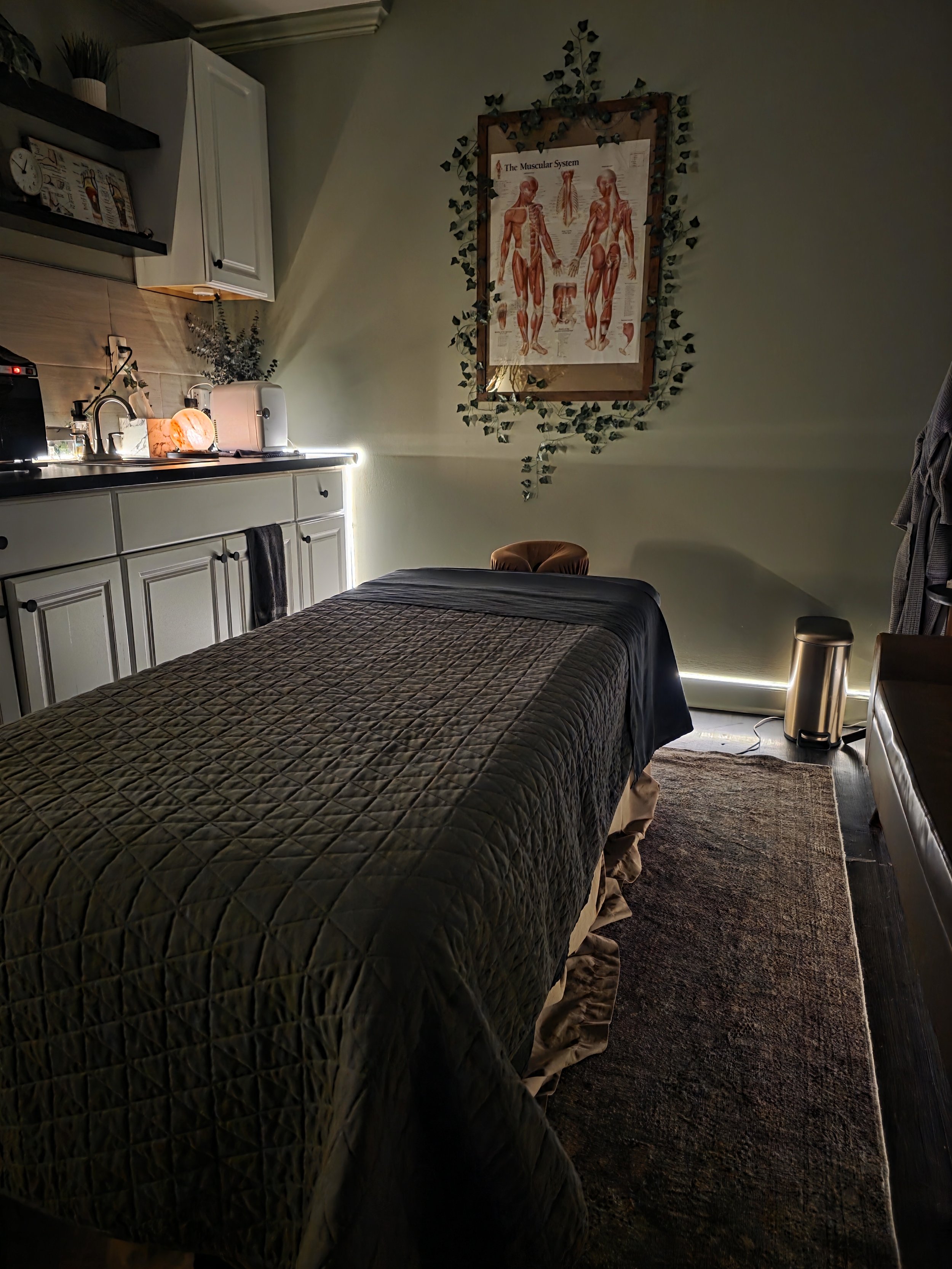 A massage or therapy room featuring a massage table with a dark quilted cover, a small fridge, a trash can, and wall decorations including a poster of the muscular system and nearby plants.