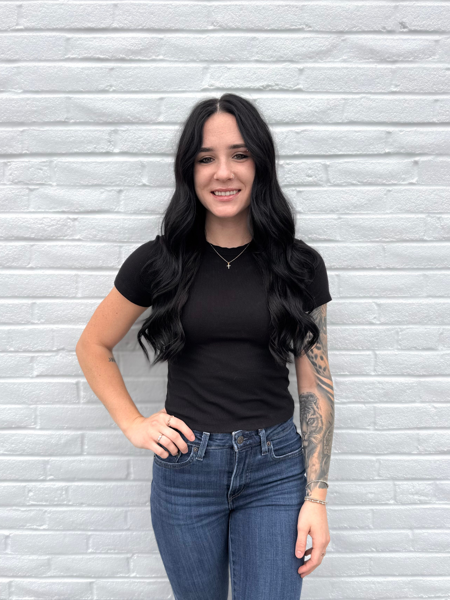 A woman with long, wavy black hair smiling at the camera. She is wearing a blue top and a necklace with cherries and crosses. She has brown eyes and a small nose piercing.