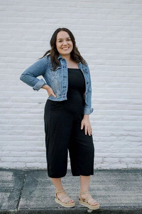 A woman with long brown hair, wearing a black jumpsuit, light blue denim jacket, and beige wedge sandals, smiling and standing against a white brick wall.