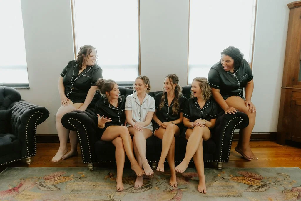 Six women in matching black pajamas sitting and standing on and around a black sofa, smiling and chatting in a room with large windows, wooden floors, and a patterned rug.