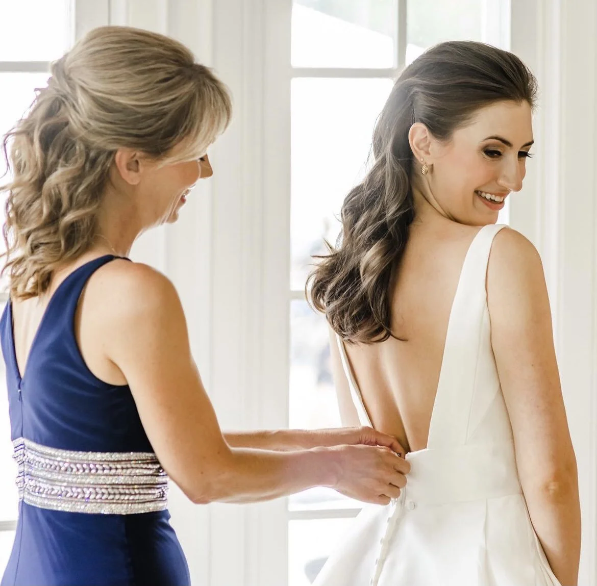 A bride in a white wedding dress smiling as an older woman helps her button the back of her dress indoors near a window.