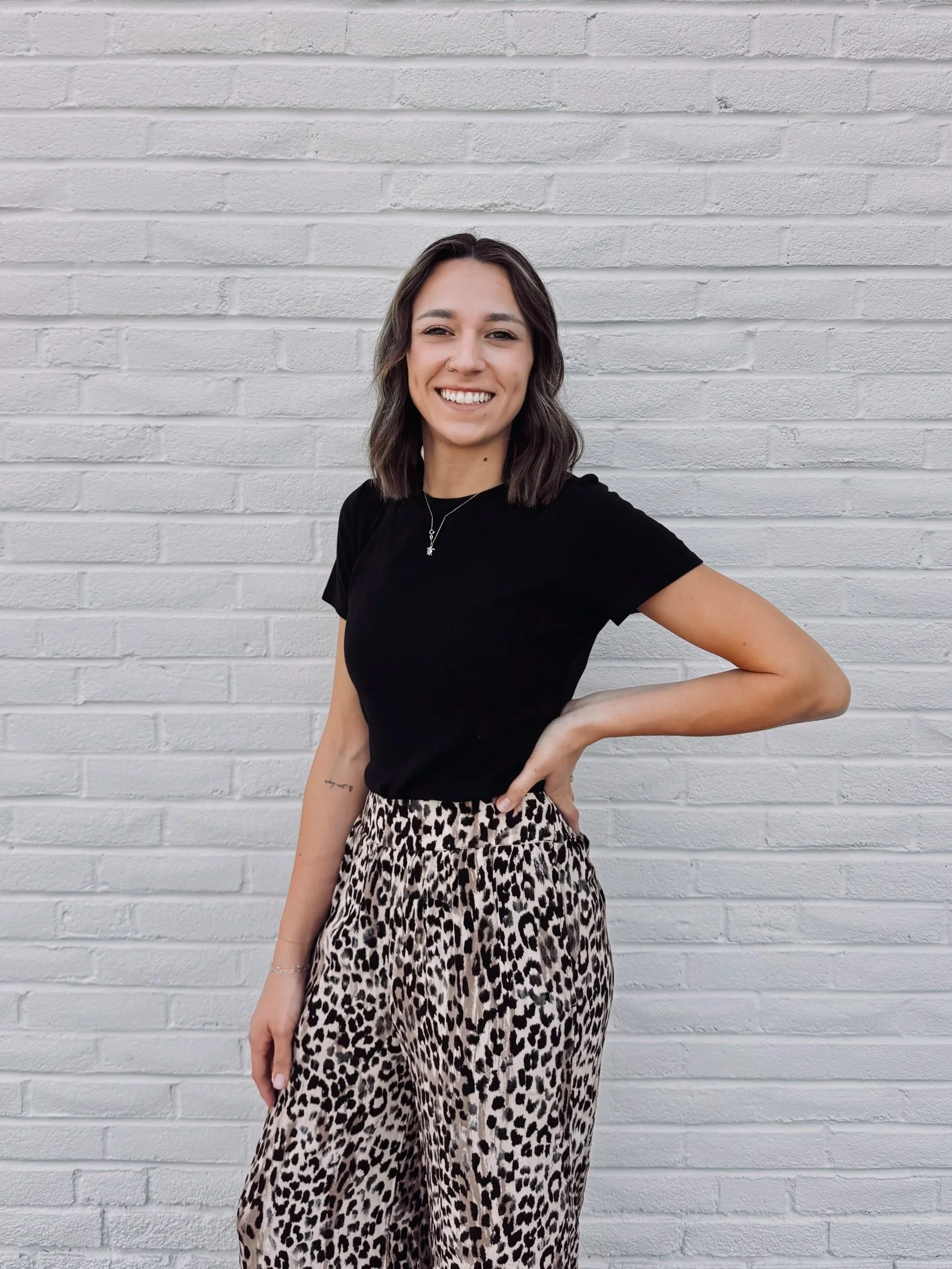 A young woman with wavy brown hair, wearing a beige sleeveless top, necklace, and black ripped jeans, smiling while standing with one hand on her hip against a plain wall.