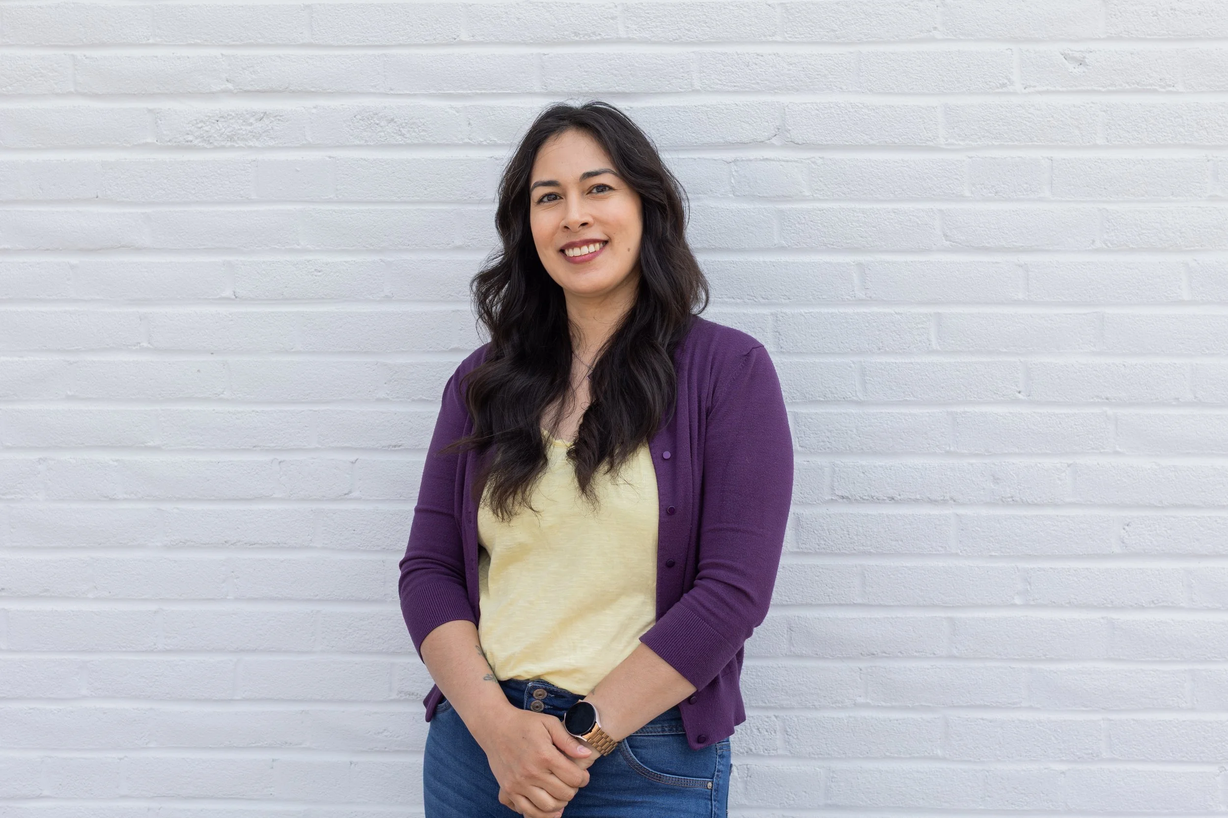 A woman with long dark hair wearing a purple cardigan, yellow top, and jeans standing in front of a white brick wall, smiling.