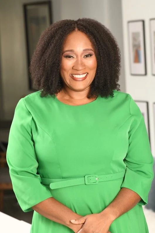 A woman with curly black hair smiling, wearing a bright green dress with three-quarter length sleeves and a matching belt, standing indoors with picture frames on the wall behind her.