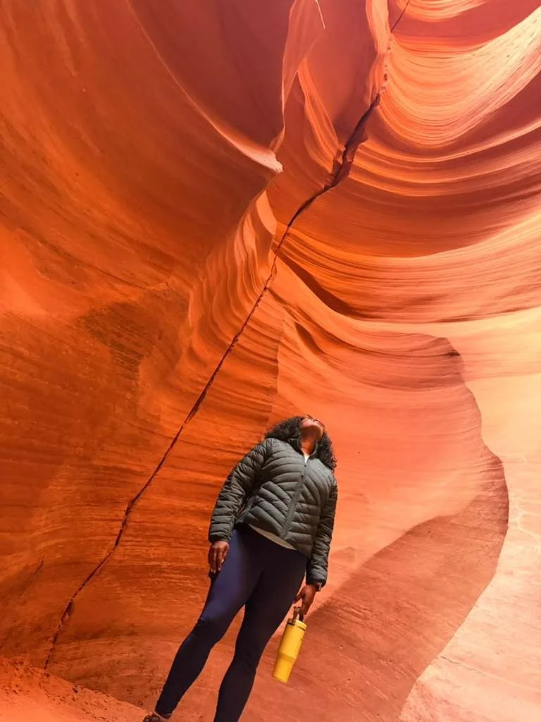 A woman standing inside a narrow, winding slot canyon with smooth, flowing sandstone walls in shades of orange, red, and tan, holding a yellow water bottle, wearing a black puffer jacket and dark leggings.