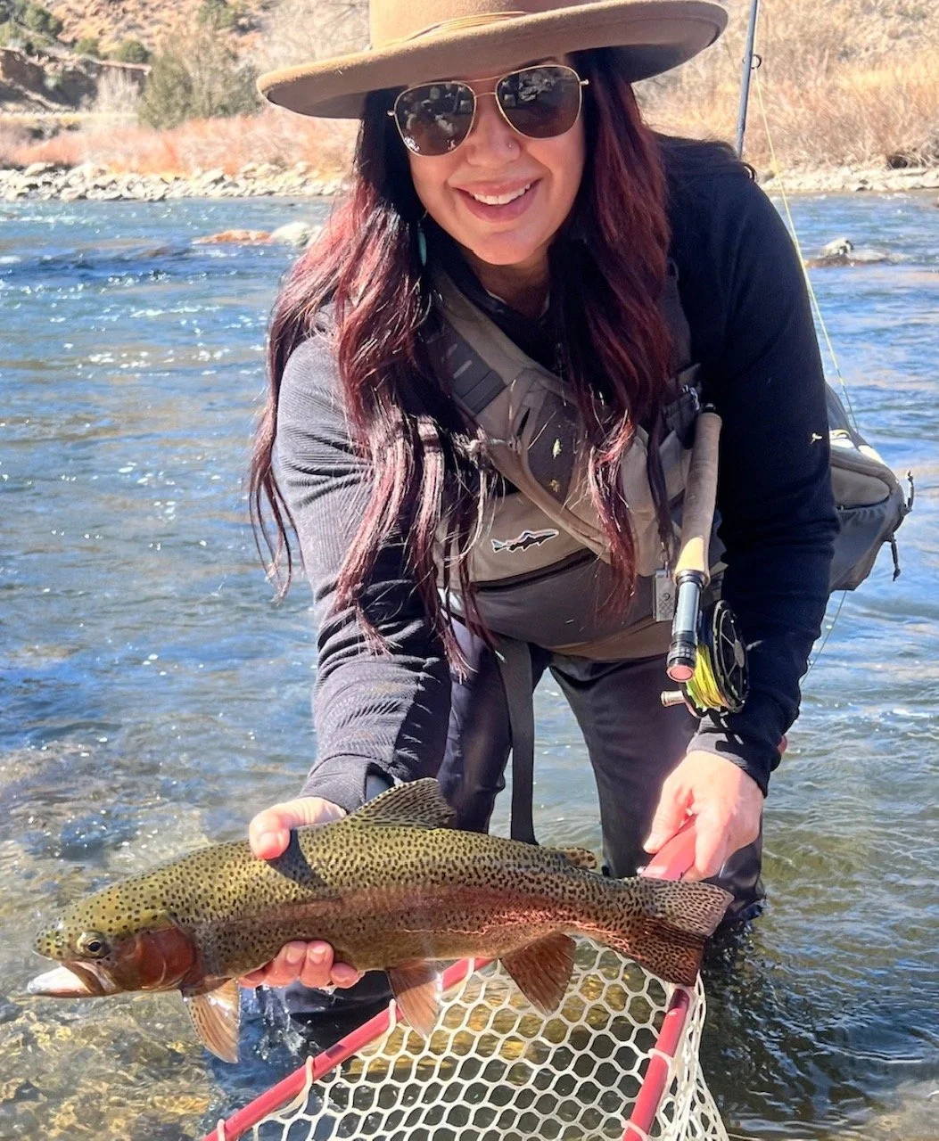 A woman with long red hair, wearing sunglasses, a hat, and fishing gear, crouching in a river holding a rainbow trout she caught.