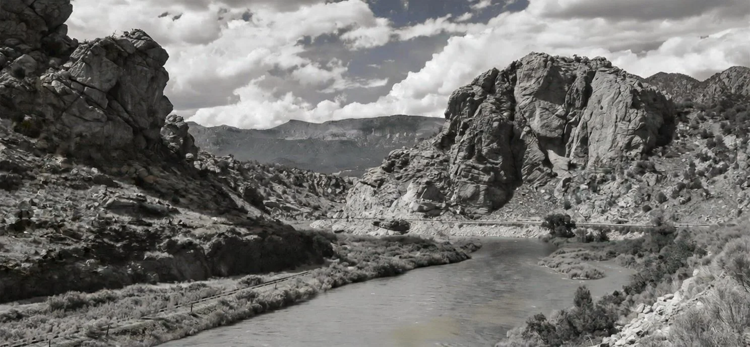 A black and white landscape photo of a river flowing through a rocky canyon with large cliffs and mountains in the background under a cloudy sky.