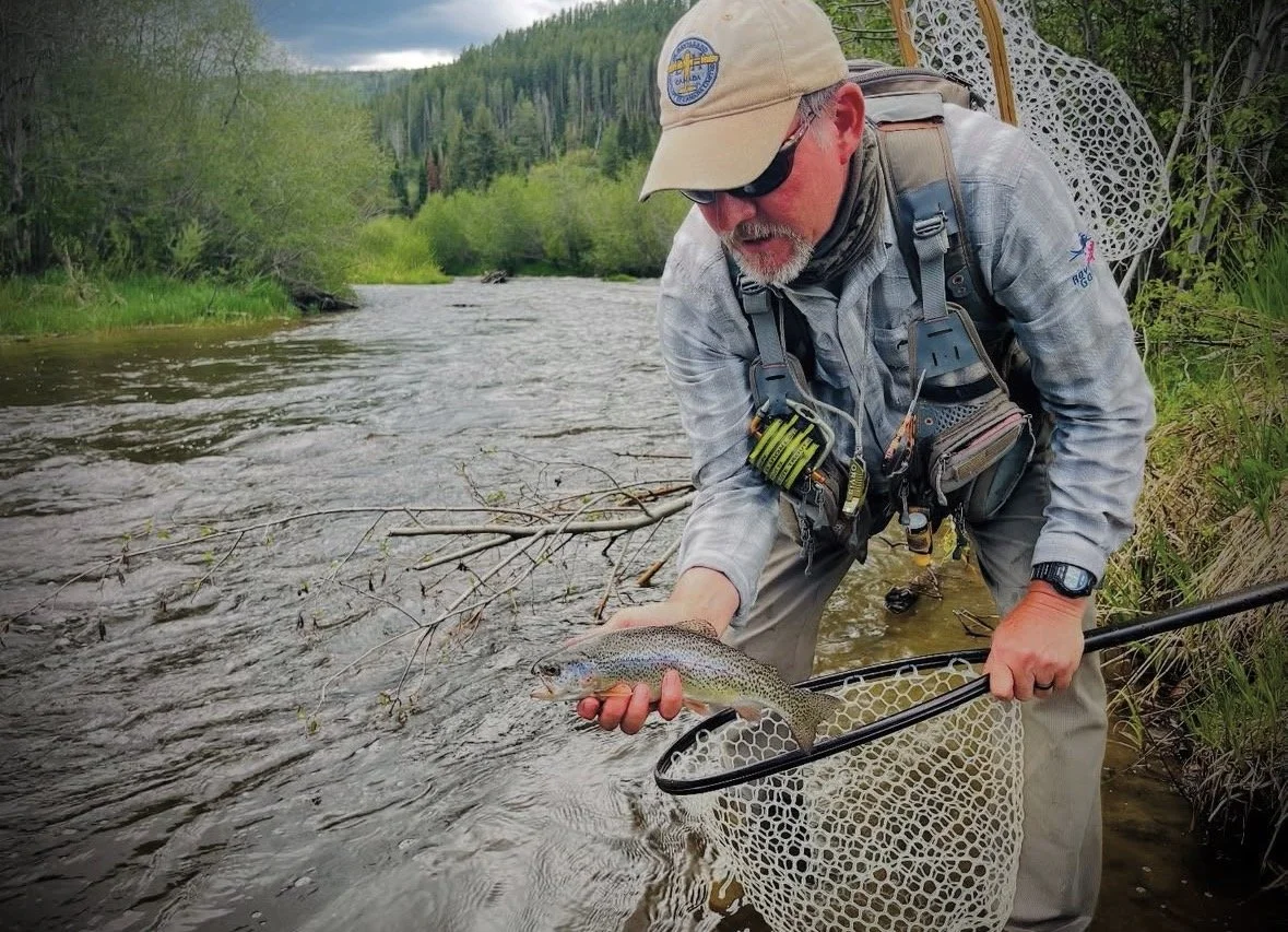 Man in fishing gear holding a rainbow trout in a river with lush green trees and mountains in the background.
