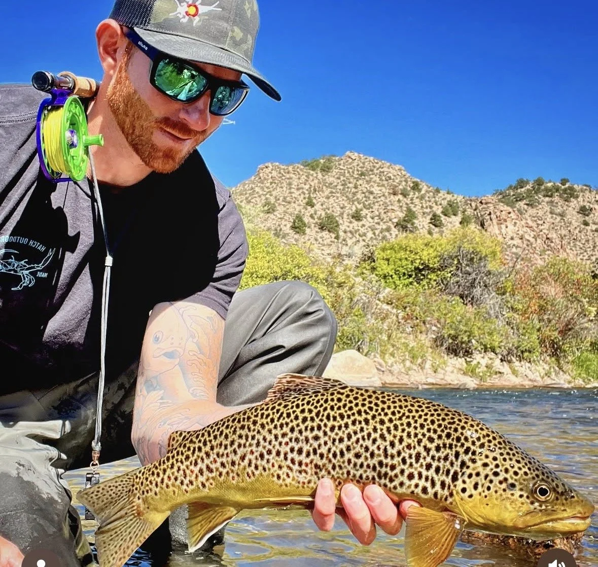 Man with tattoos and sunglasses holding a spotted fish by the river, with mountains and trees in the background.