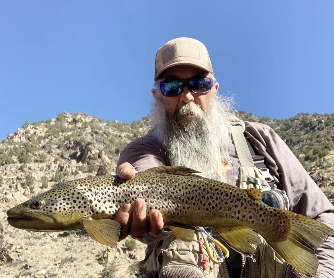 A man with a long gray beard wearing sunglasses and a tan cap holding a large fish outdoors, with a mountainous background and clear blue sky.
