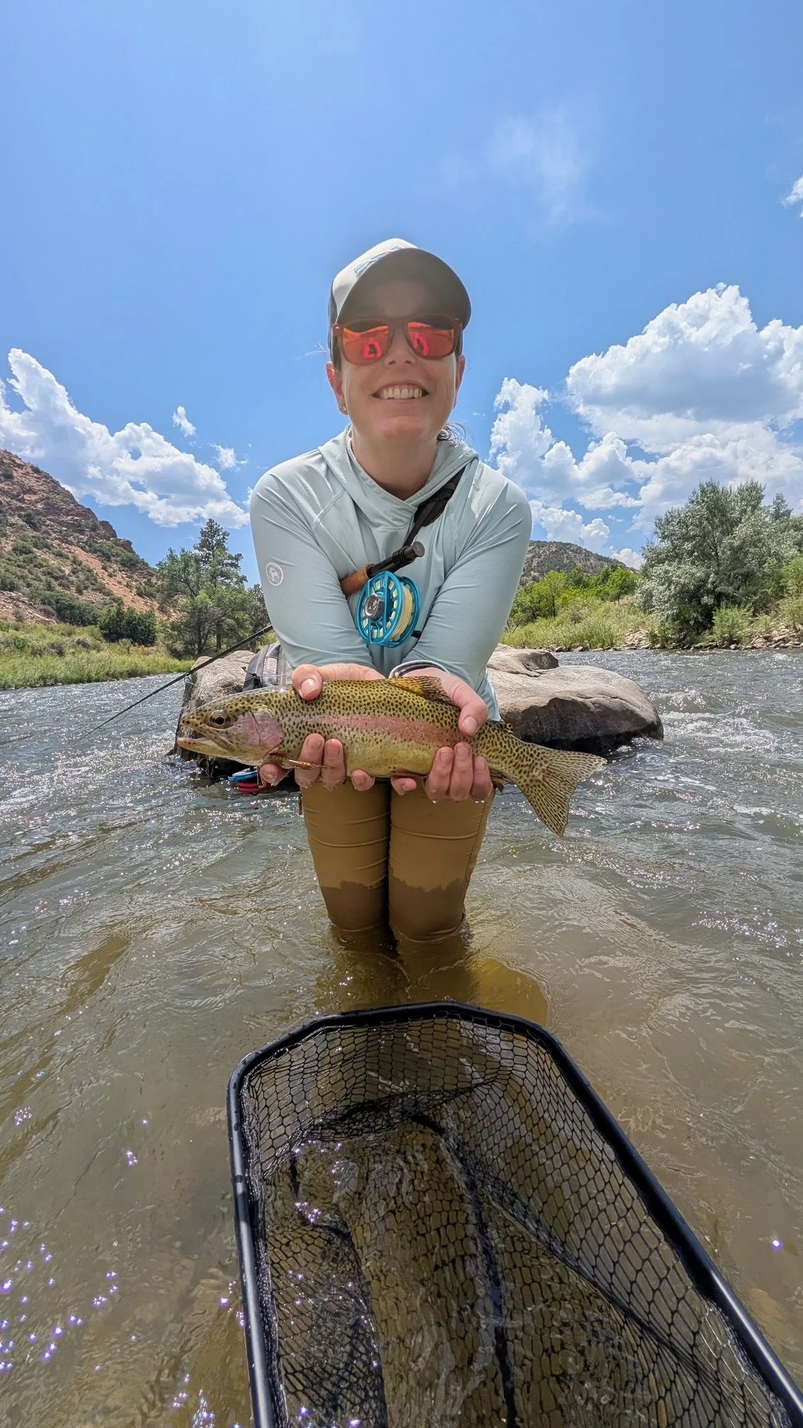Person standing in a river holding a large rainbow trout, wearing fishing gear and sunglasses, with a scenic background of mountains and cloudy sky.