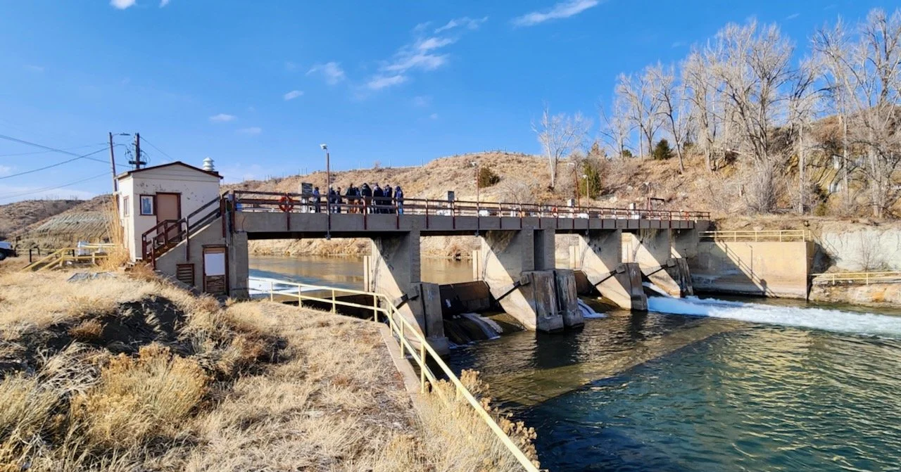 A small hydroelectric dam with water flowing through it, surrounded by dry grass and leafless trees, under a clear blue sky.