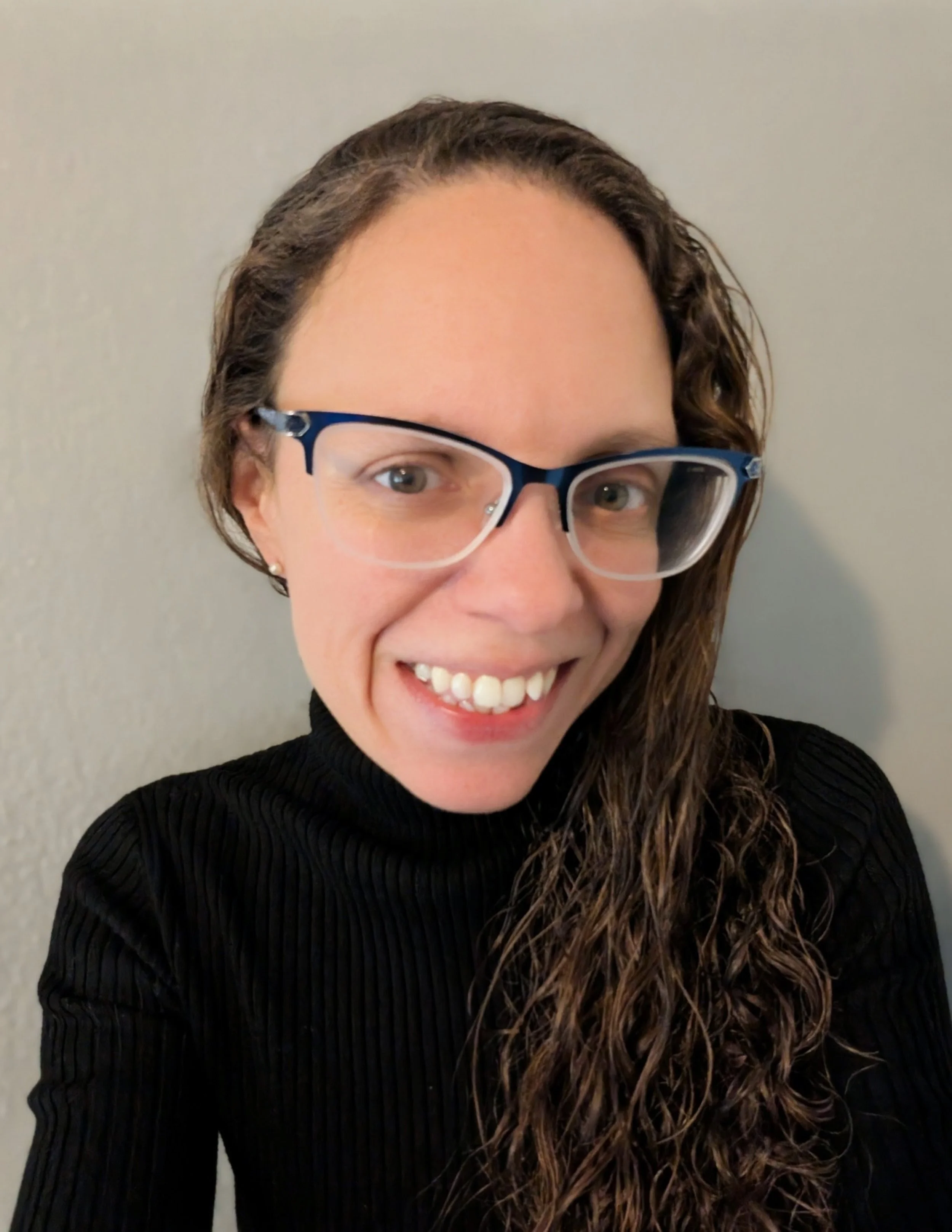 Close-up photo of a woman with curly brown hair, wearing glasses with dark blue frames and a black ribbed turtleneck sweater, smiling at the camera against a plain light gray background.