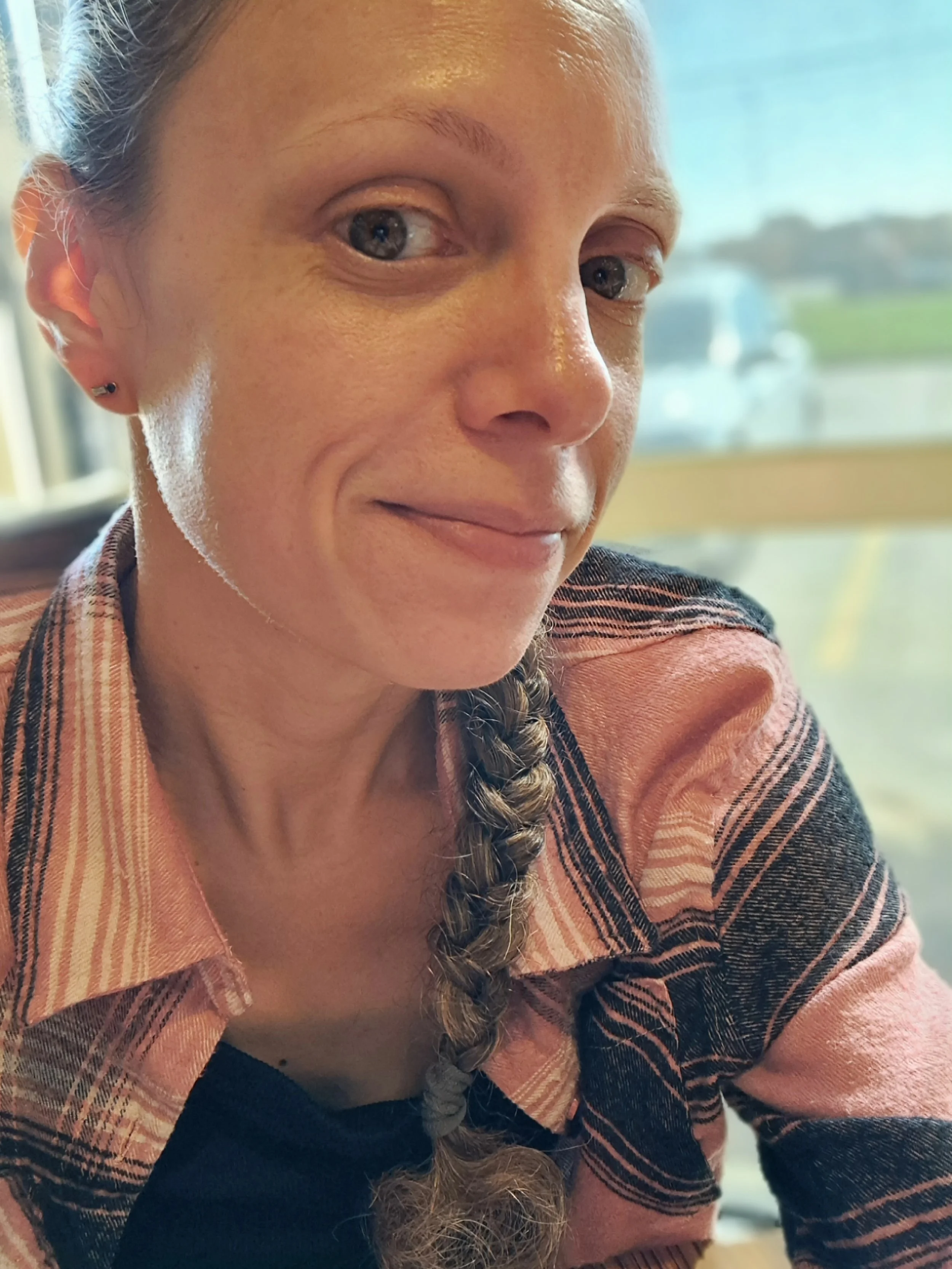 A woman with braided hair and small earrings sitting indoors, with a blurry outdoor scene visible through the window behind her.