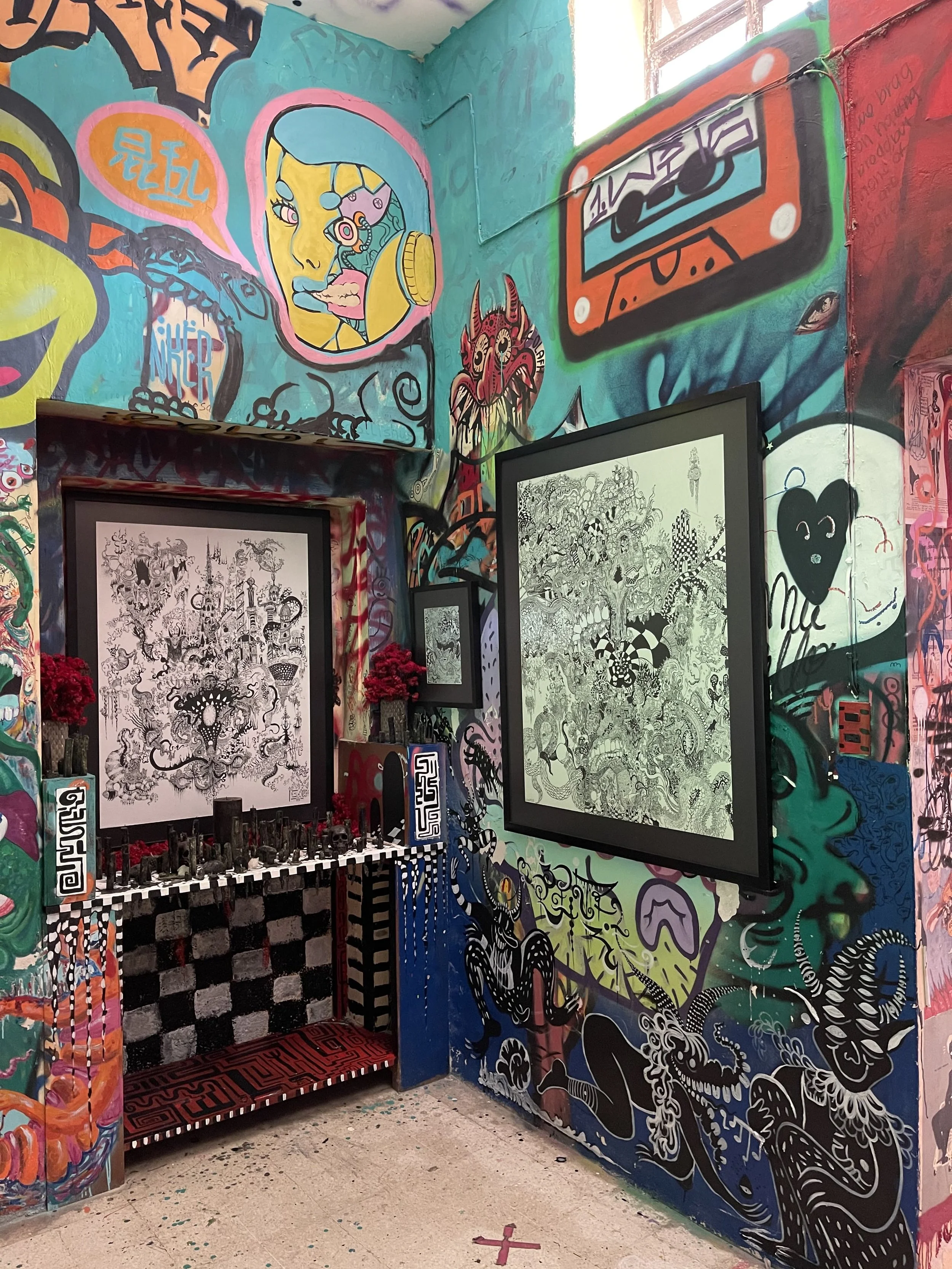 Habitación decorada con arte callejero y grafiti, con obras enmarcadas en la pared, una mesa de negras y blancas, y letras en lo alto. Muchas piezas de arte, desde dibujos en blanco y negro hasta colores vivos, cubren las paredes y el techo.