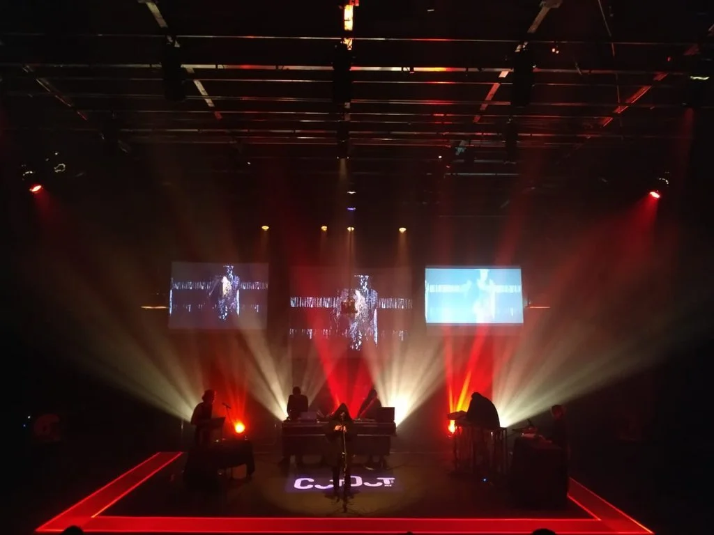 Escenario con músicos en vivo, iluminación dramática en tonos rojos y amarillos, pantallas con imágenes en el fondo.