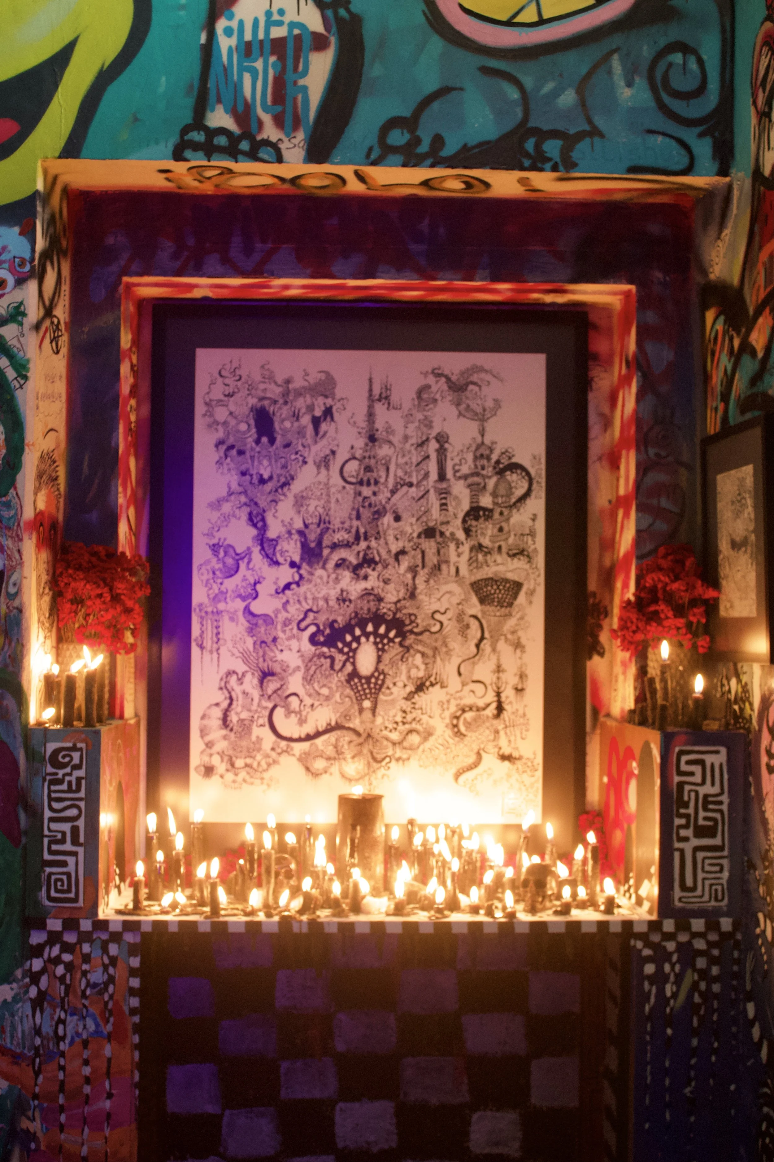 Un altar decorado con muchas velas encendidas, flores rojas y un cuadro con un dibujo detallado en el fondo, en medio de un mural colorido y artístico.