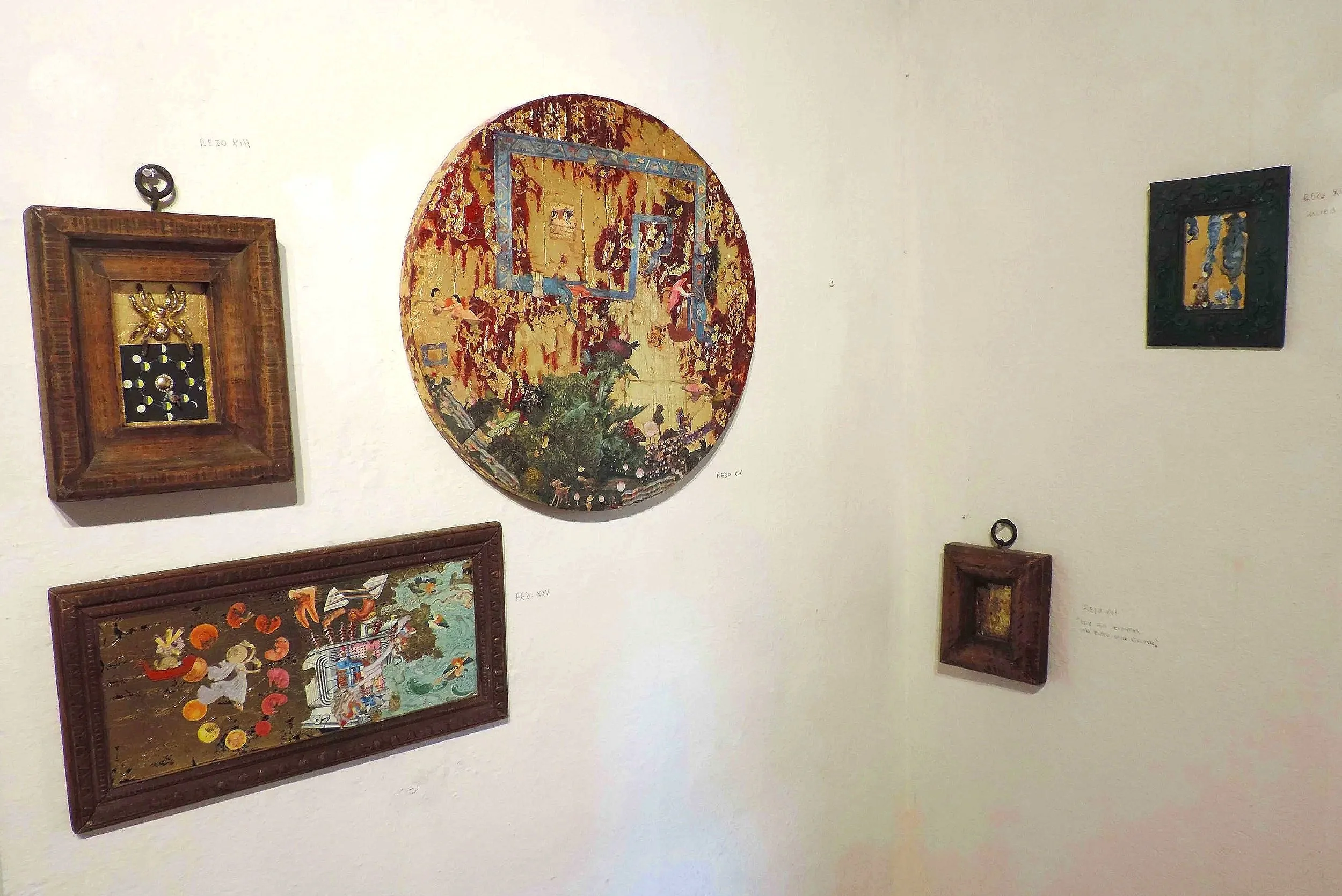 Diversas obras de arte enmarcadas y un cuadro circular en una pared blanca. Algunas obras tienen estilos abstractos y figurativos.