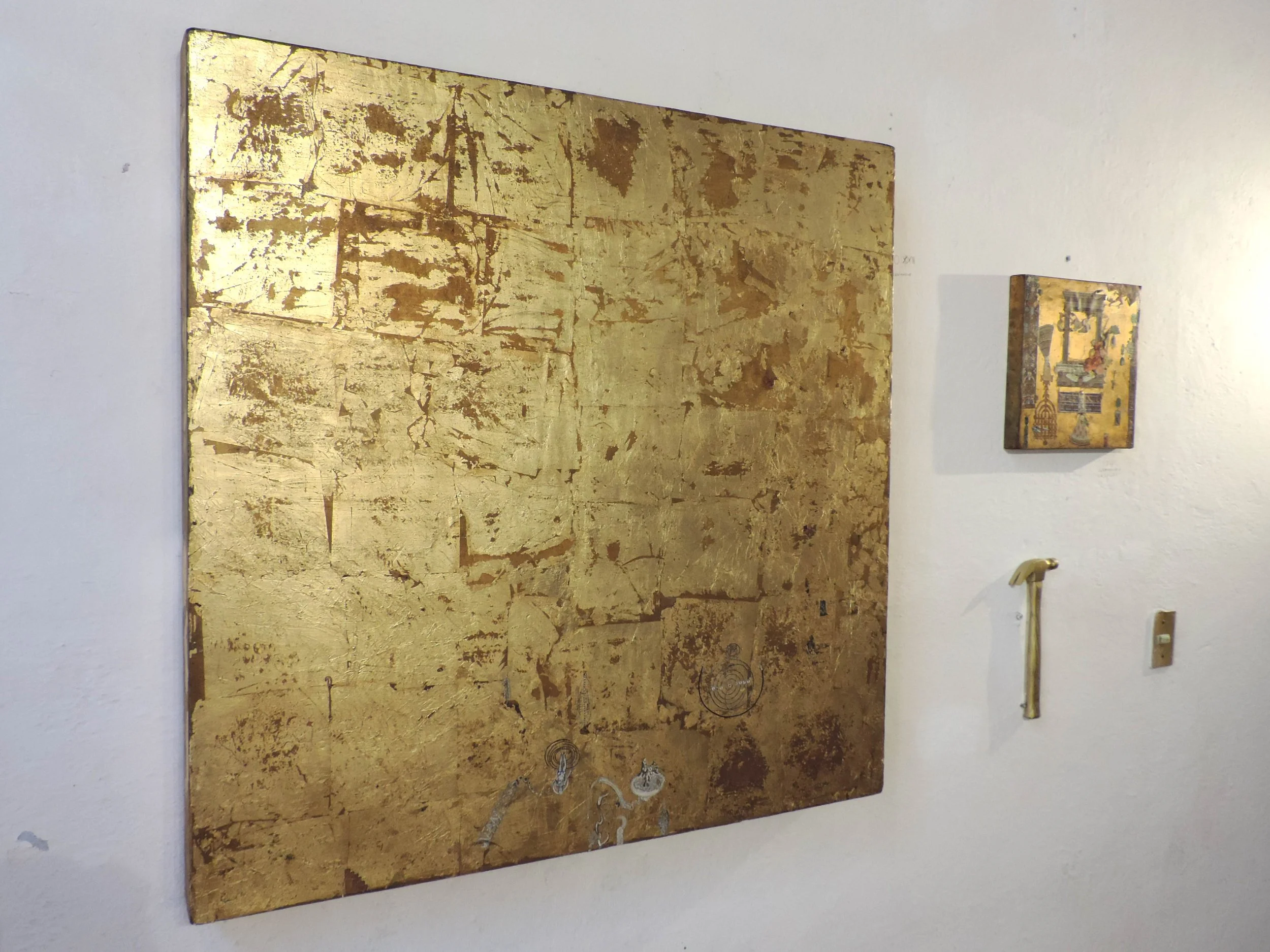 Pintura abstracta con fondo dorado y detalles en plateado, y piezas pequeñas colgadas en la pared blanca.