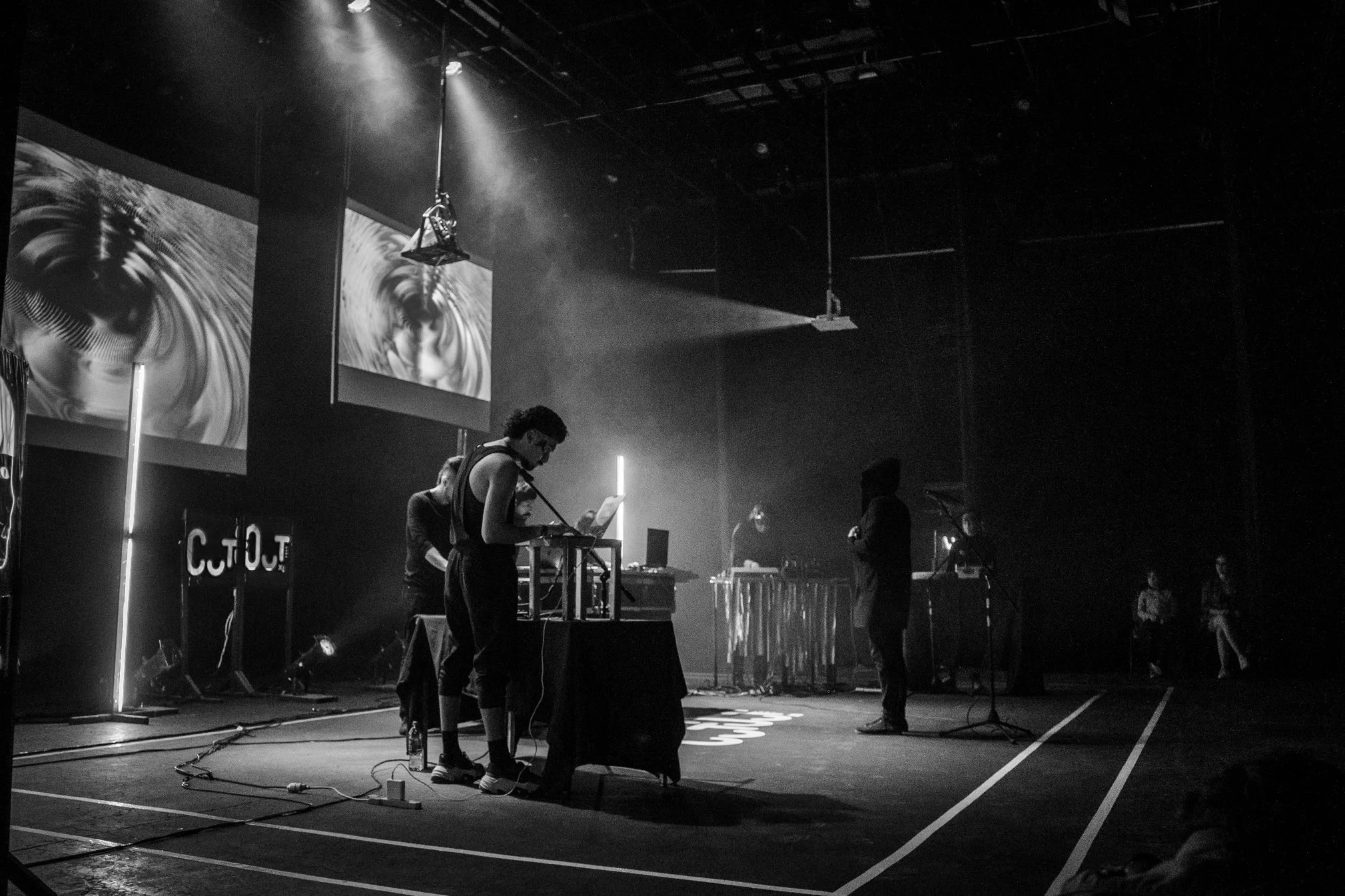 Escenario oscuro con músicos y equipo de sonido, pantallas de fondo con visuales abstractos, algunos espectadores sentados en la esquina.