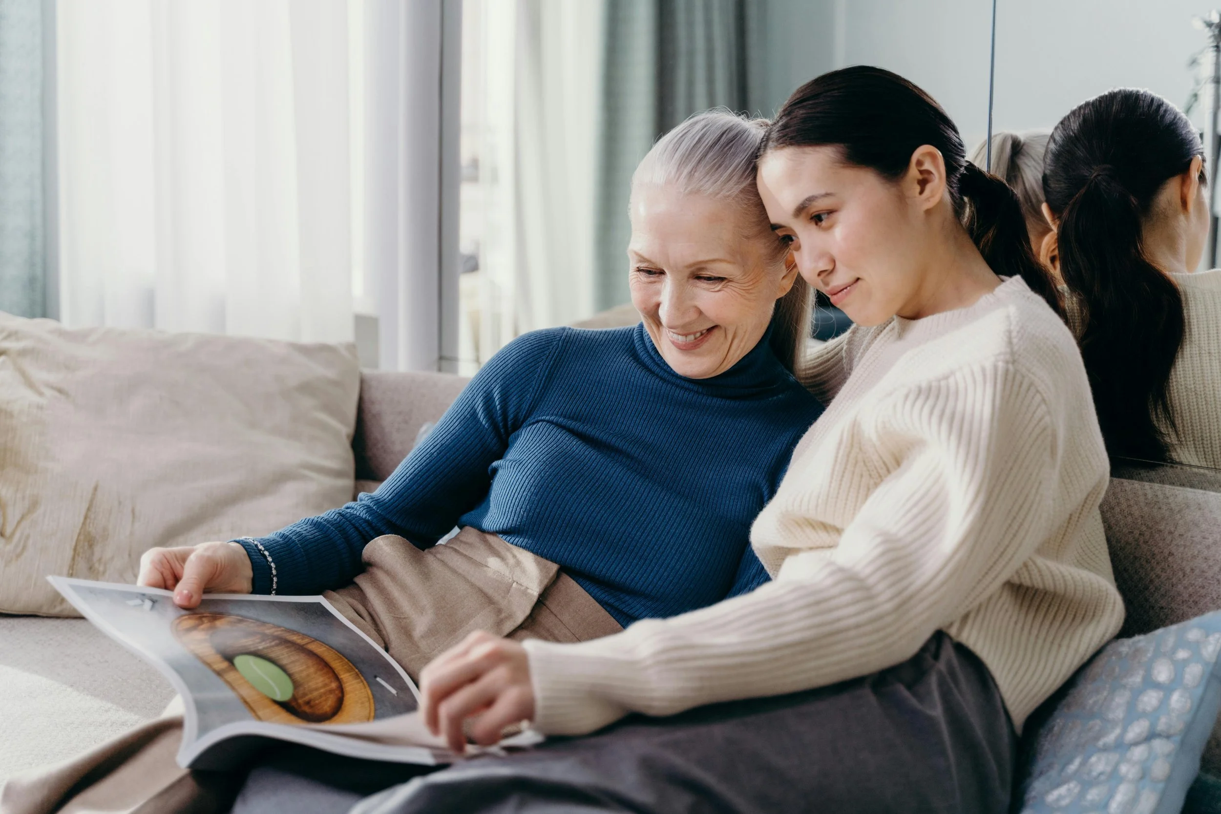 Two women, one elderly with gray hair and one younger with dark hair, sitting close on a sofa, looking at an open magazine together, smiling.