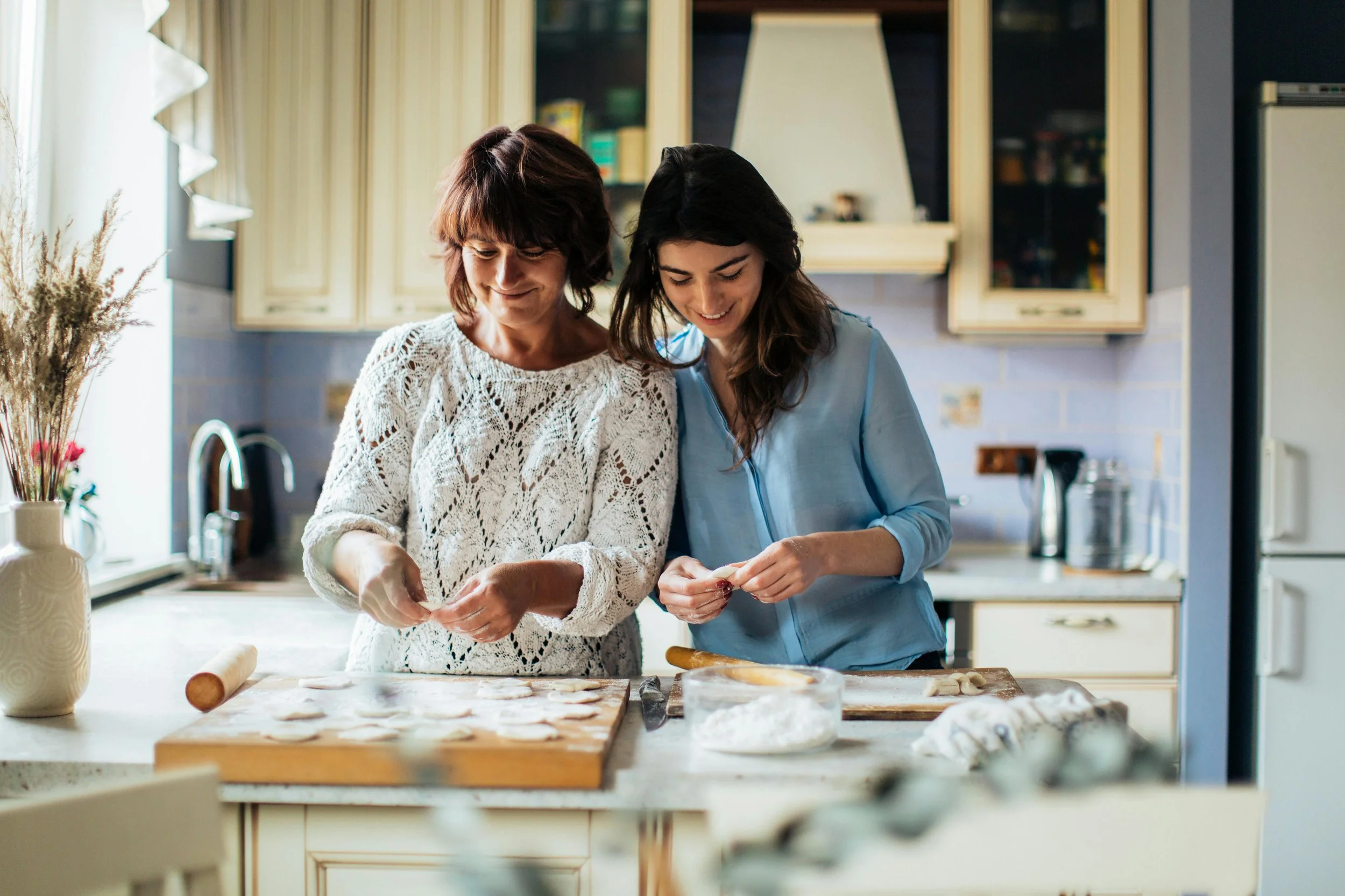 Two women baking cookies together in a bright kitchen, smiling and working on dough on a wooden countertop.