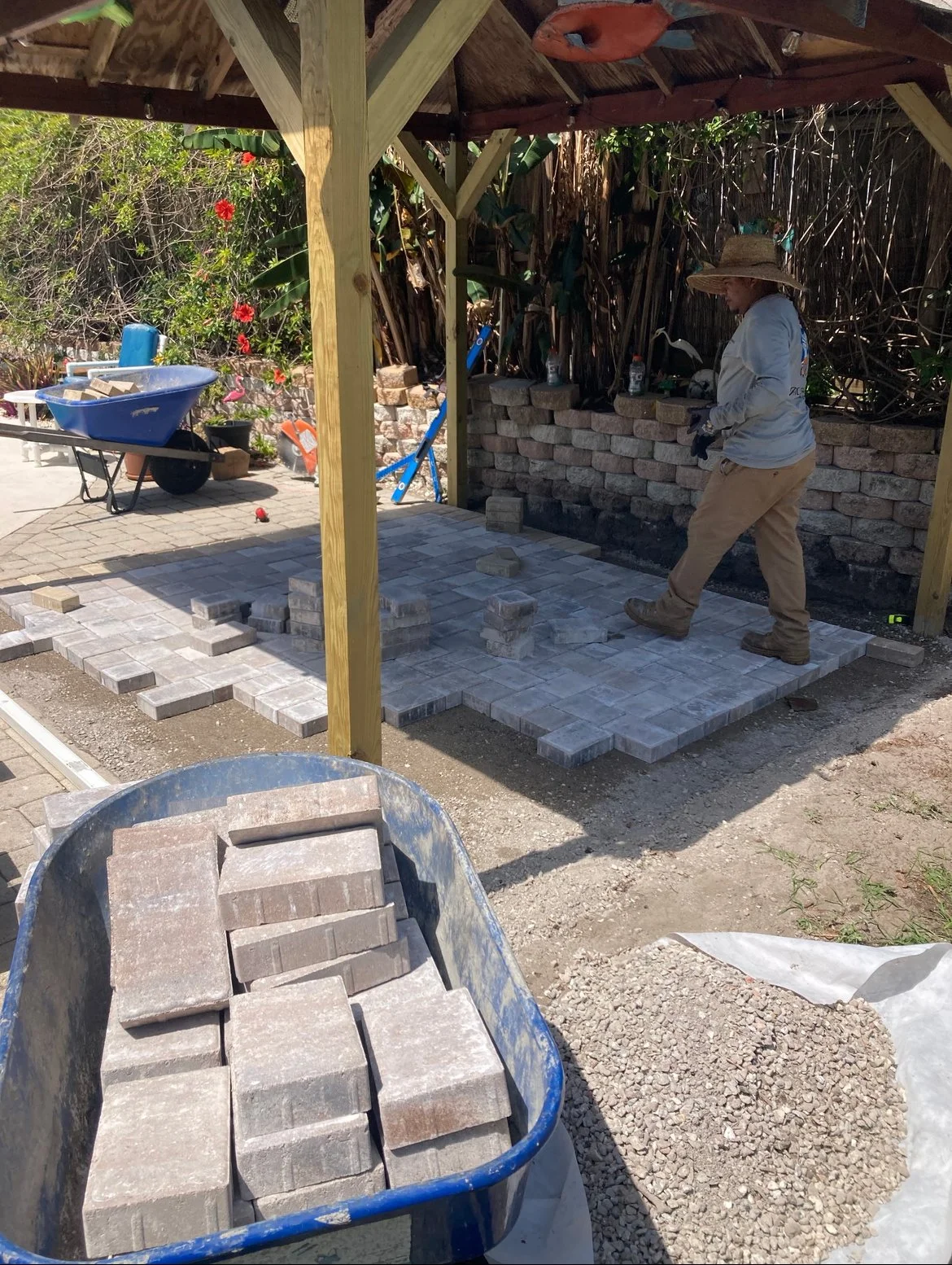 A person working outdoors laying paving stones on a patio under a wooden structure. Various tools and materials, including a wheelbarrow full of bricks, are visible around the work area.