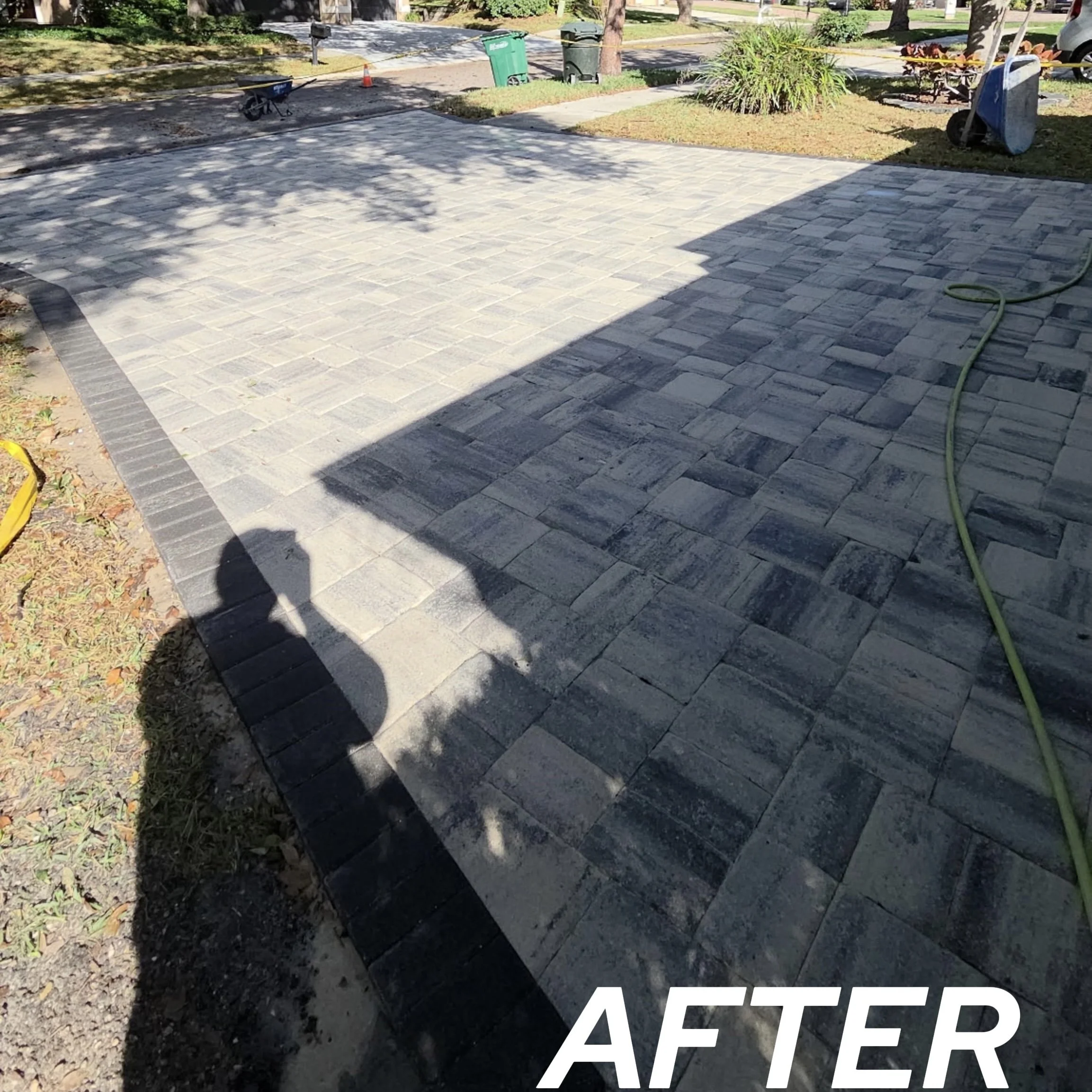 Freshly installed gray and black stone brick patio with a hose on the right side and shadows of trees and a person taking the photo.