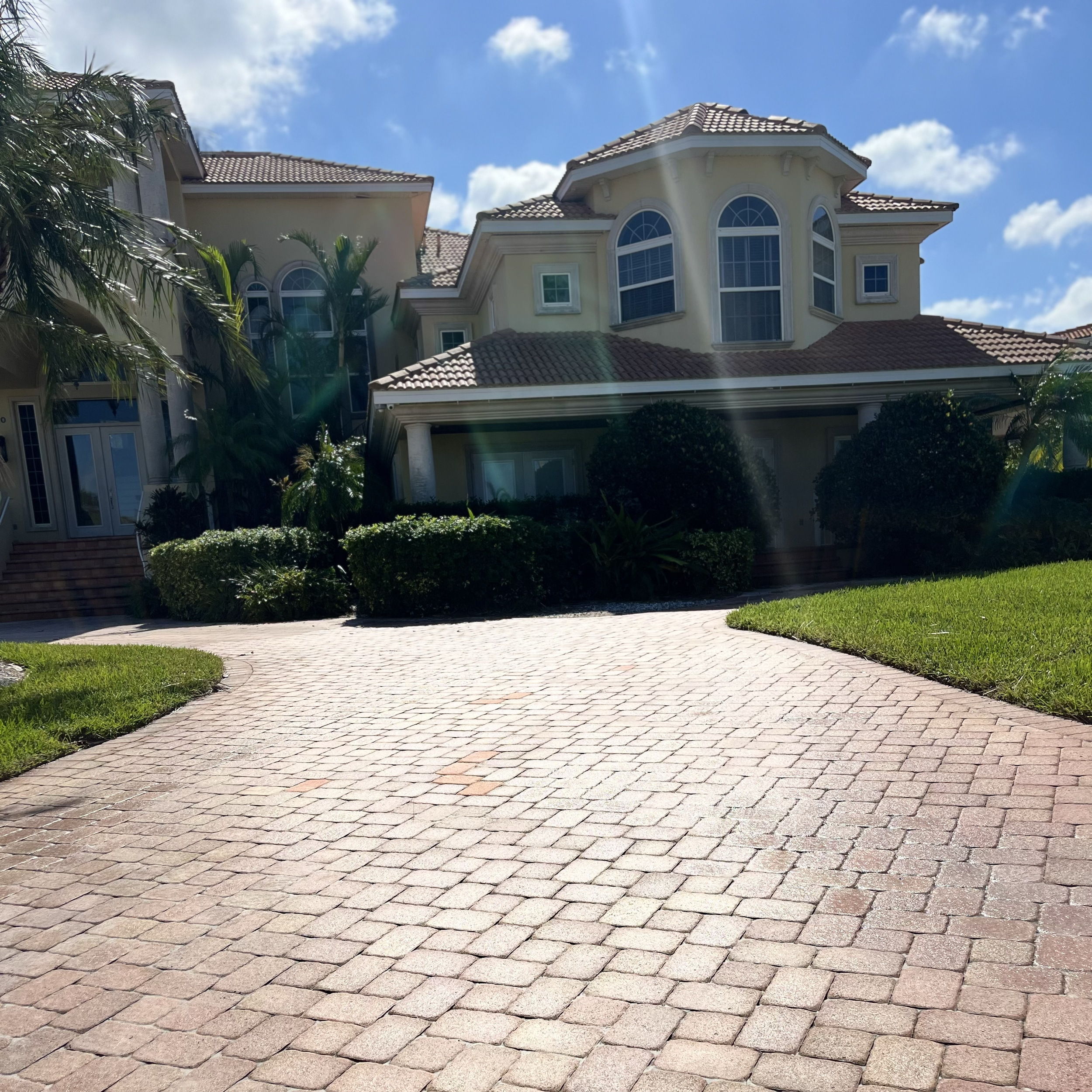 Front view of a large, elegant house with a paved driveway, lush green grass, and tropical plants under a partly cloudy blue sky.