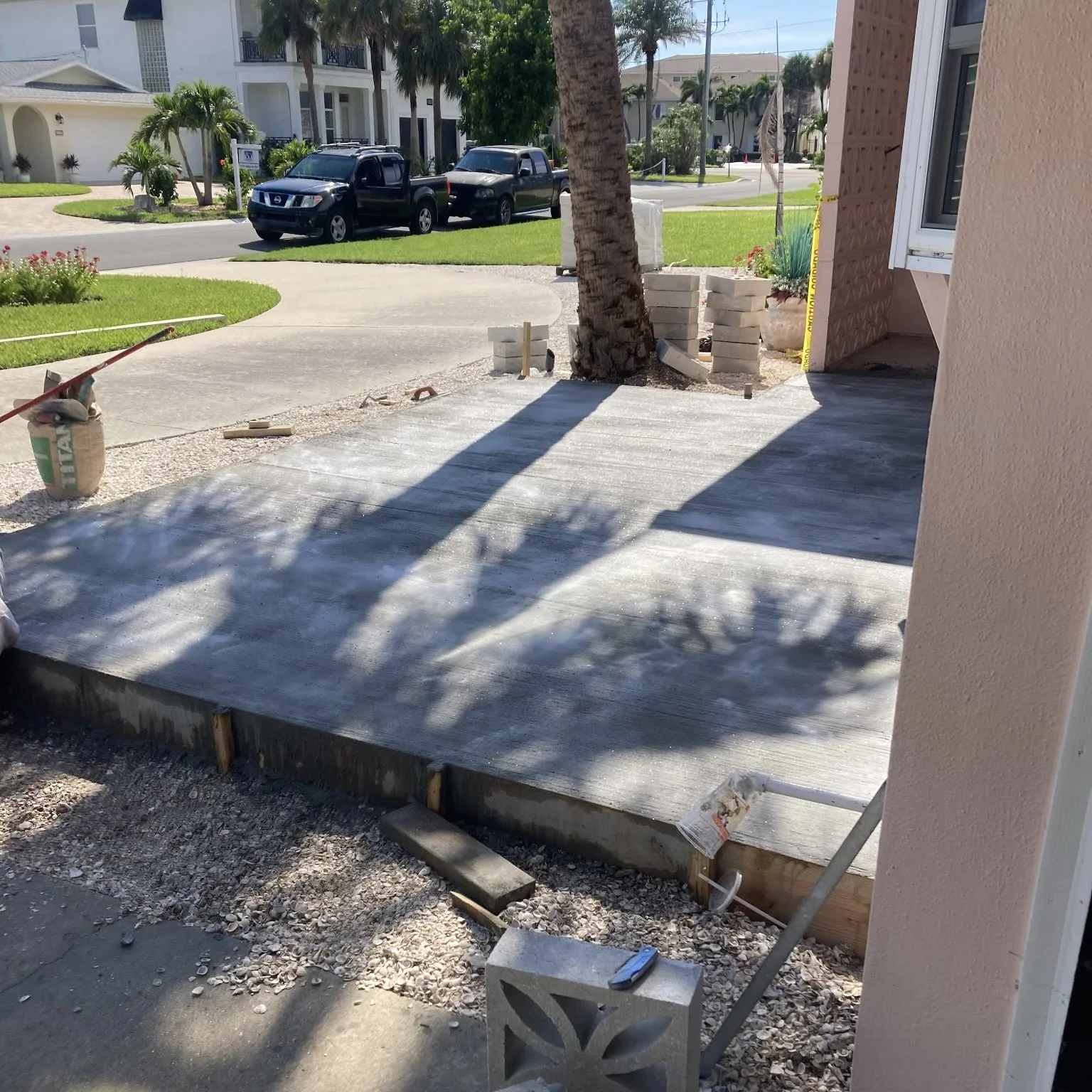 Construction of a concrete patio in a residential area with construction materials and tools, including a trowel and cinder blocks, while shadows of palm trees are cast on the surface.