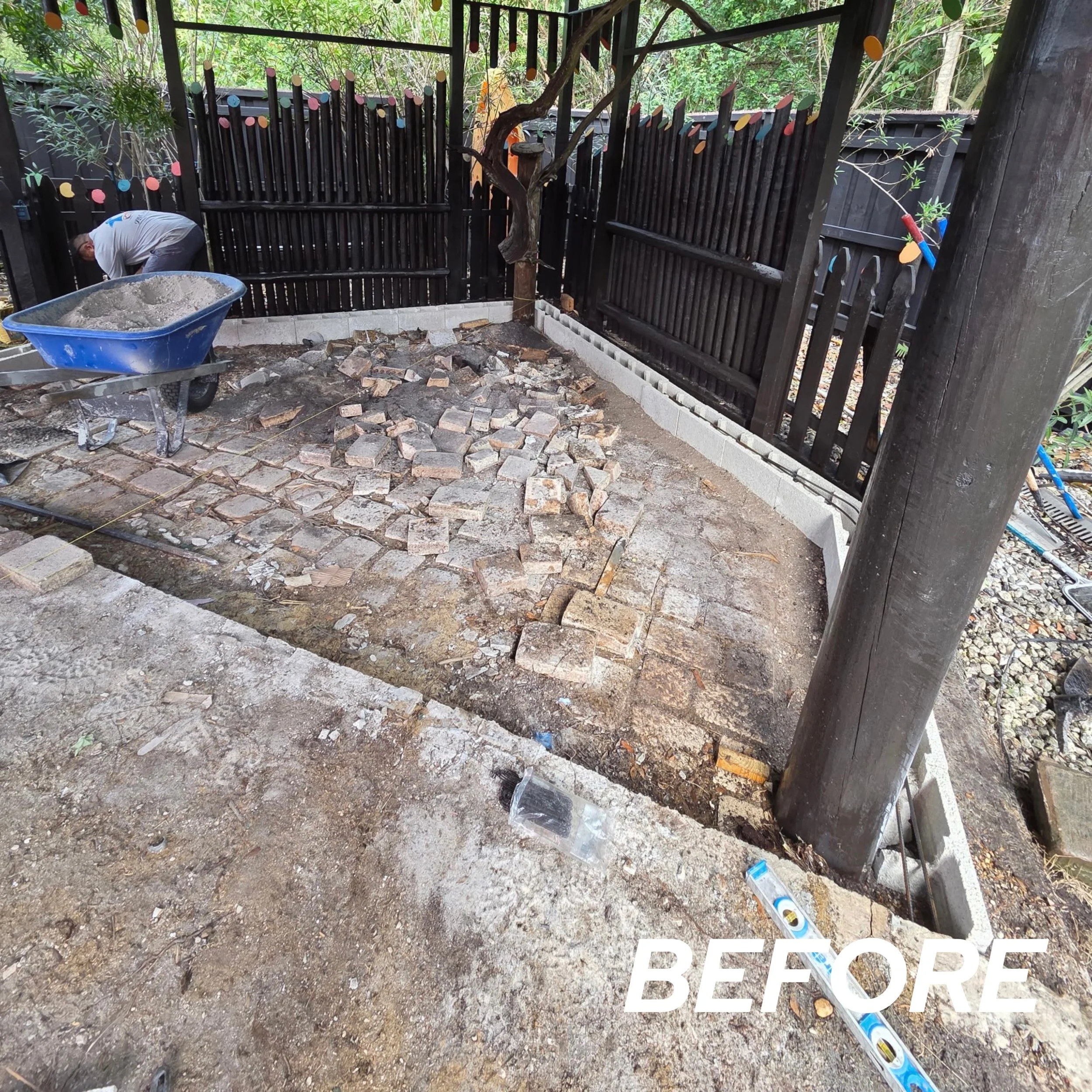 Backyard under renovation with partially laid brick patio, wheelbarrow filled with sand, and a worker in the background. Fencing and a tree are visible.