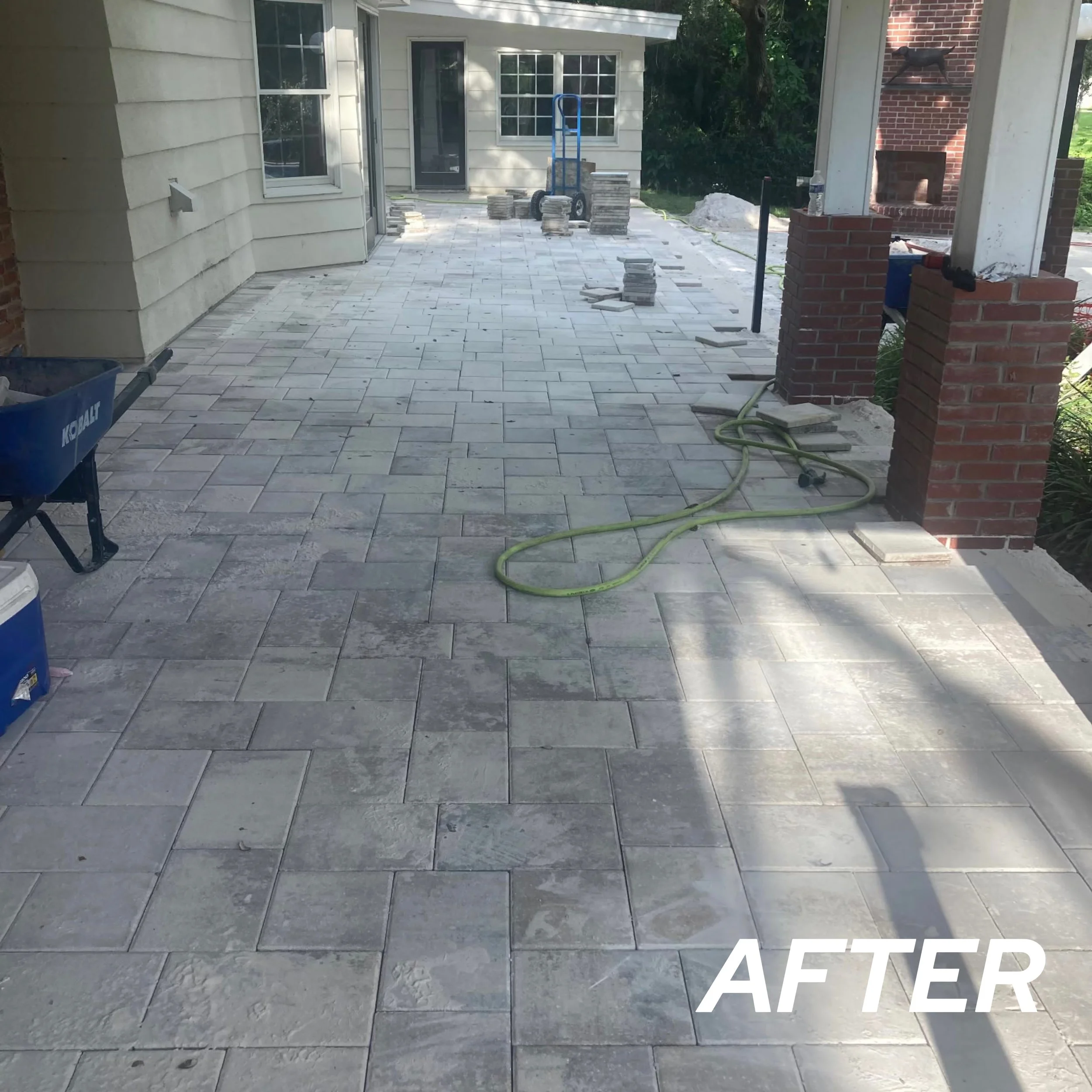 Newly installed stone patio on a porch with a yellow hose, construction tools, and stacked paving stones. The porch features brick columns and a white clapboard house.