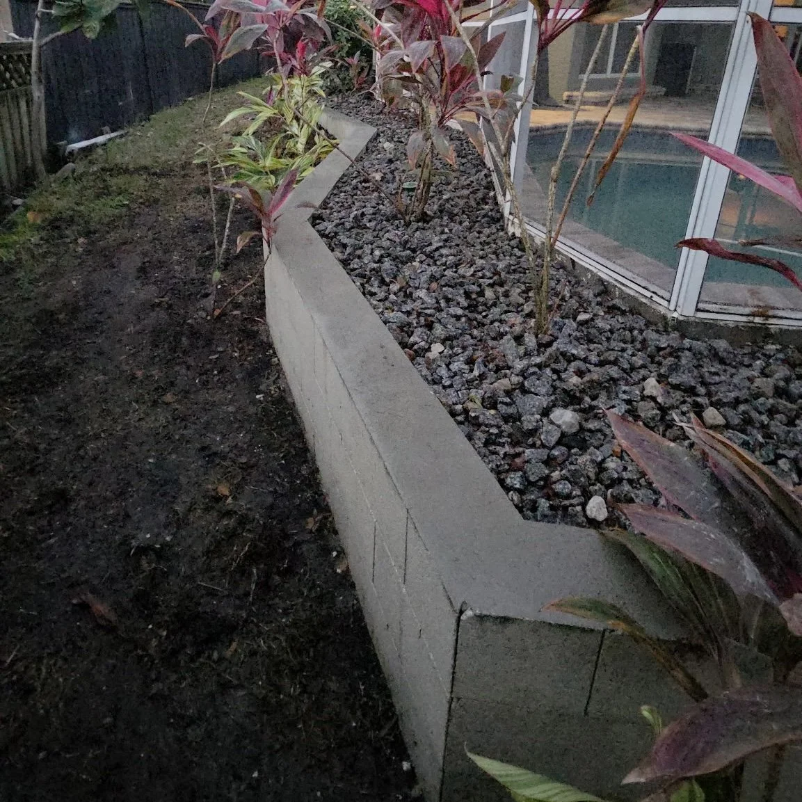 A garden bed made of concrete blocks, filled with small rocks and plants with reddish leaves, located next to a glass enclosed porch or sunroom.
