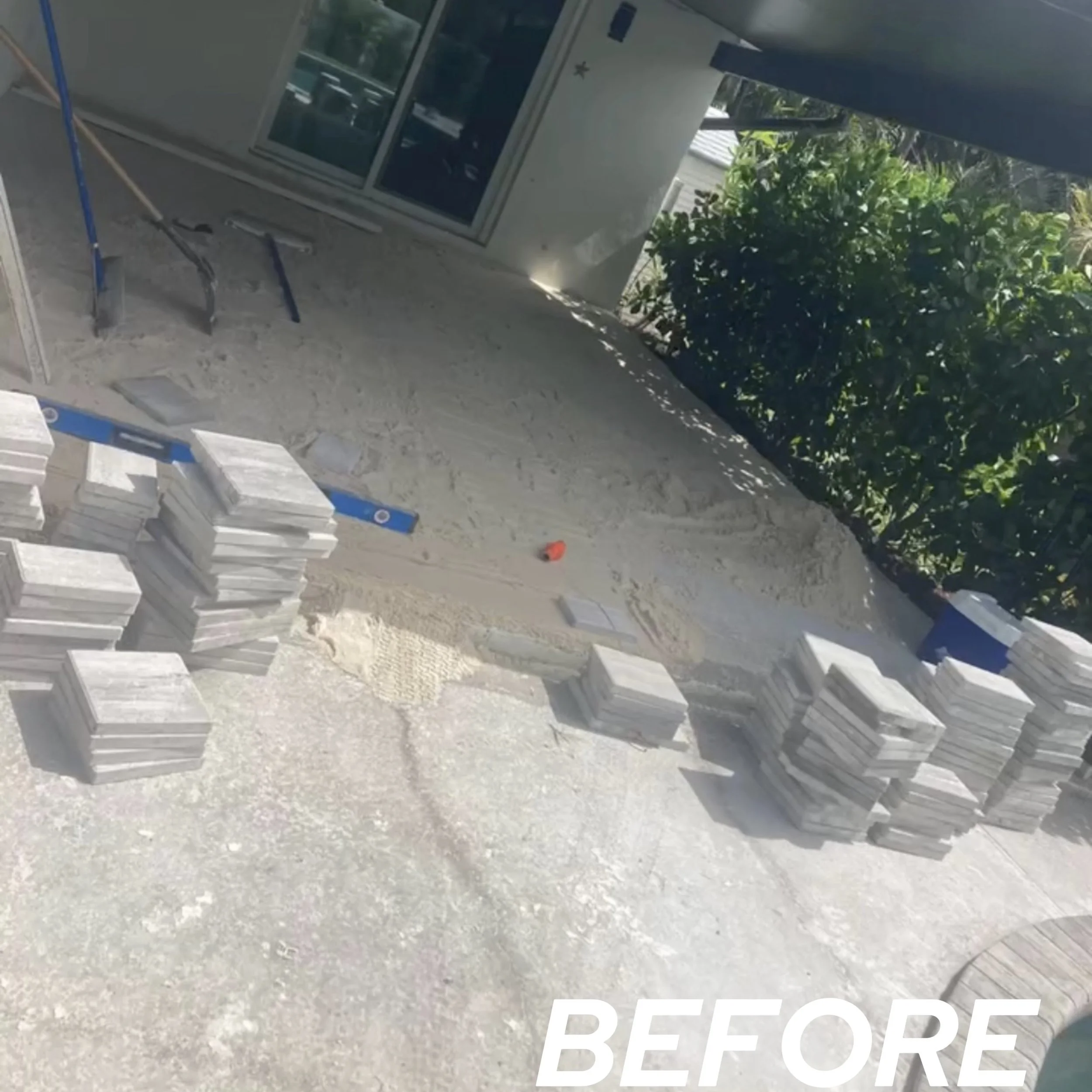 Stacks of tiles and construction tools arranged outside a house under renovation, with a window and green shrubbery in the background.