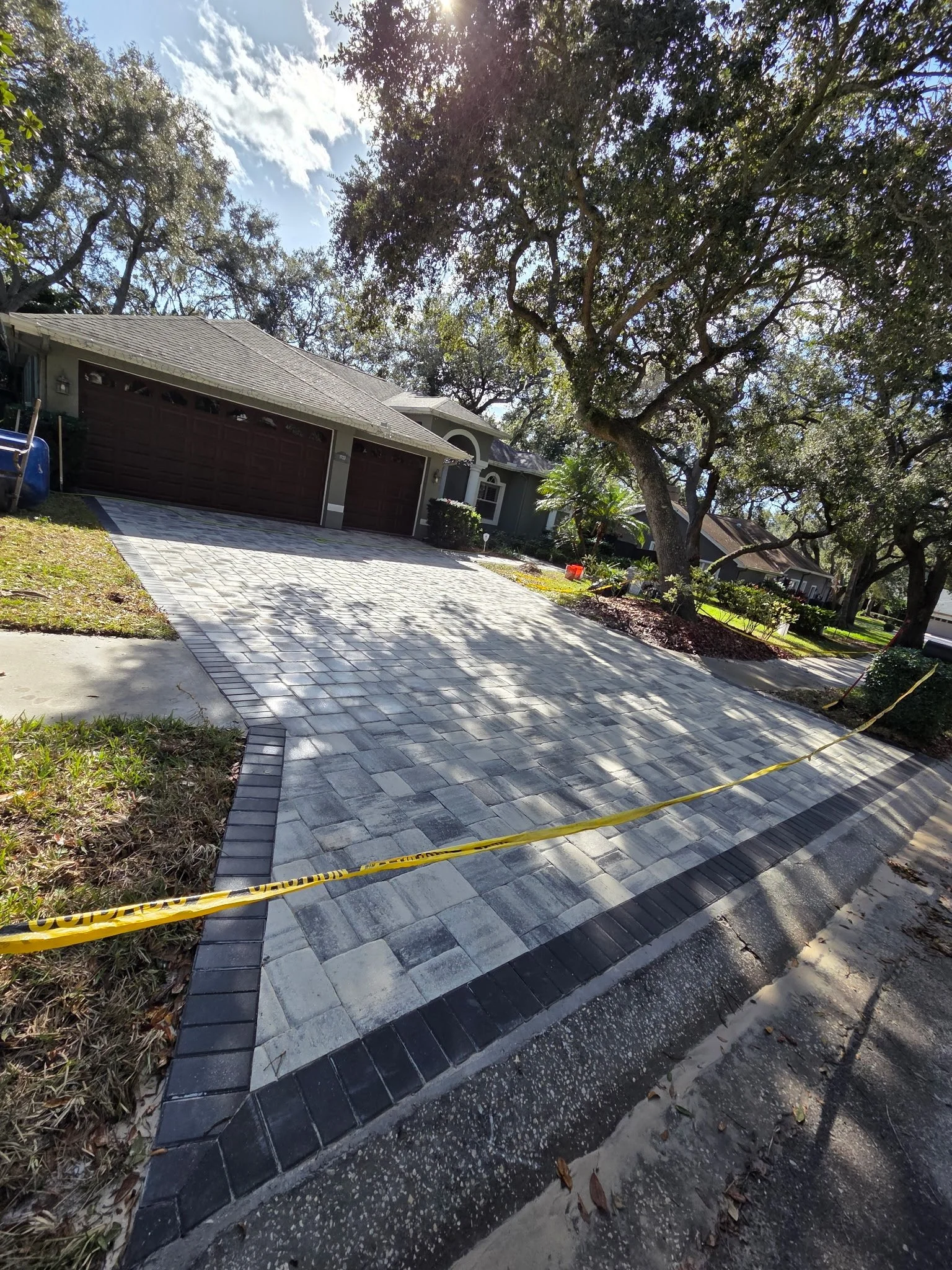 Newly paved driveway in front of a house with a two-car garage, large trees, and a front yard with bushes, under a partly cloudy sky.