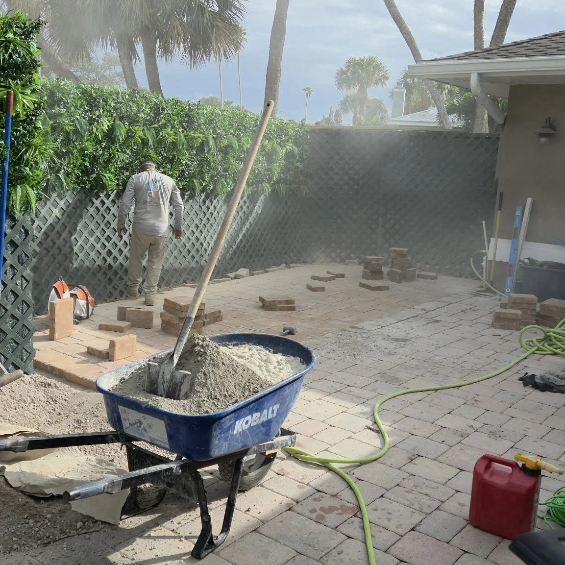 A construction worker installing paving stones on a patio, with a wheelbarrow filled with sand, stacks of bricks around, and some tools and hoses nearby. There is a lush green hedge and tall palm trees in the background.