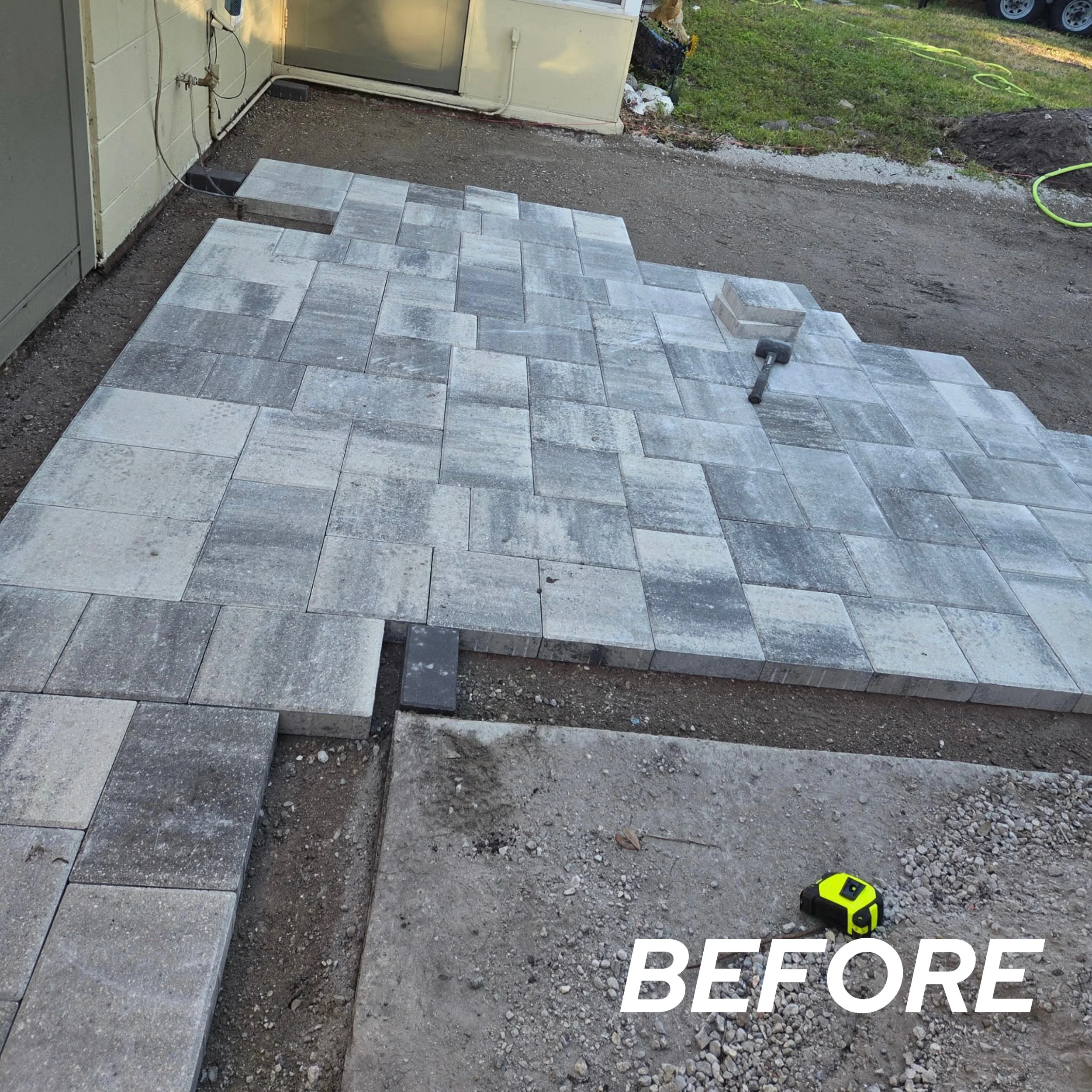 Outdoor patio area with grey stone pavers, some unevenly placed, and construction tools including a rubber mallet and measuring tape. The surrounding ground is dirt and gravel, with grass and a garden in the background. The word 'BEFORE' is written i
